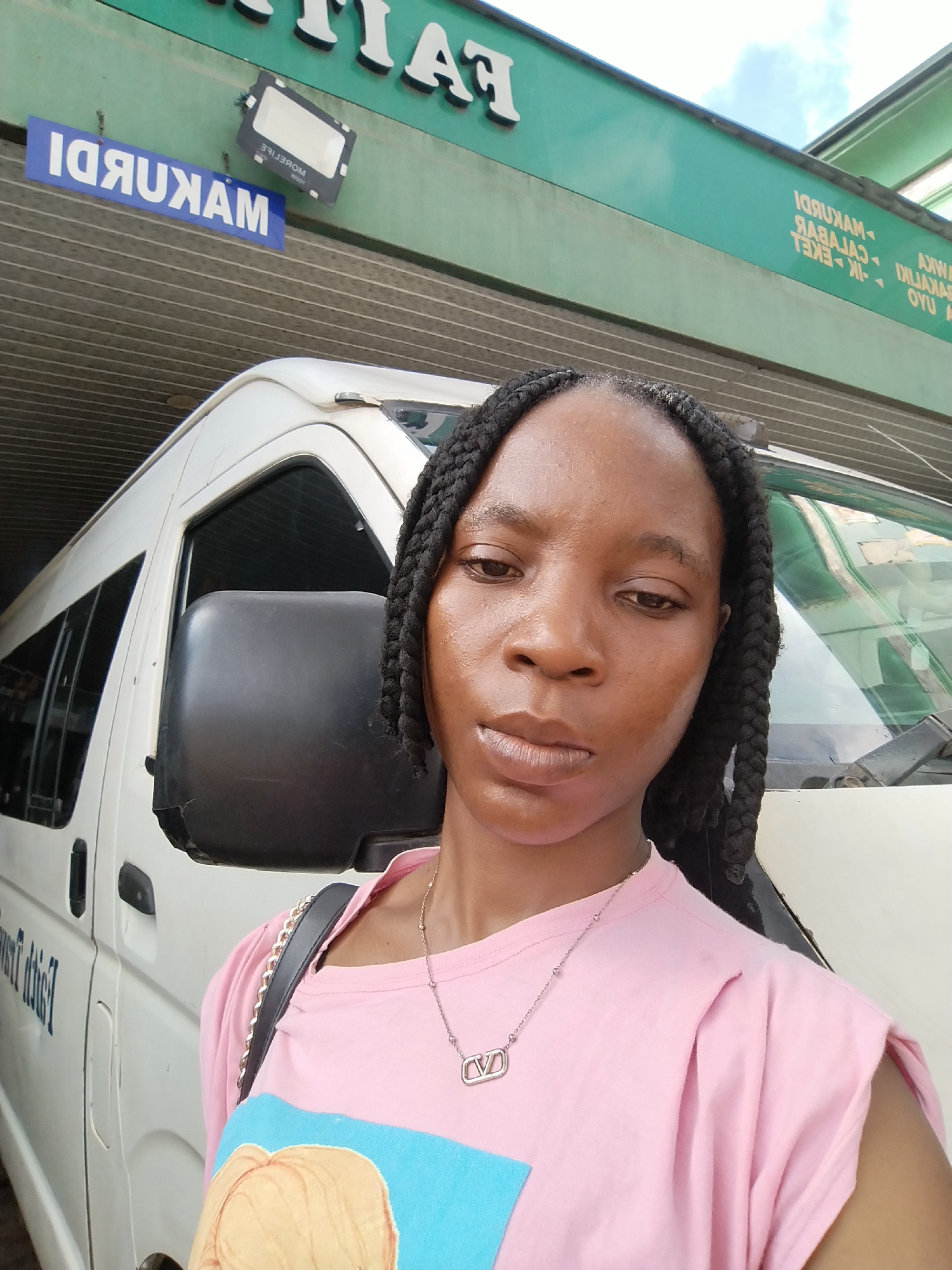 A young woman with dark skin and braided hair is taking a selfie in front of a white van. She is wearing a pink t-shirt with a graphic design and a silver necklace. The van is parked under a green awning, and there is a sign with blue lettering on the awning that reads "MAKURDI" in reverse. Another sign above the awning has white lettering that also appears to be in reverse, possibly indicating it's a company name. The location is in Benin City, Nigeria, likely a bus station or transport hub, suggested by the presence of the van and signage indicating destinations. The lighting suggests it is daytime.