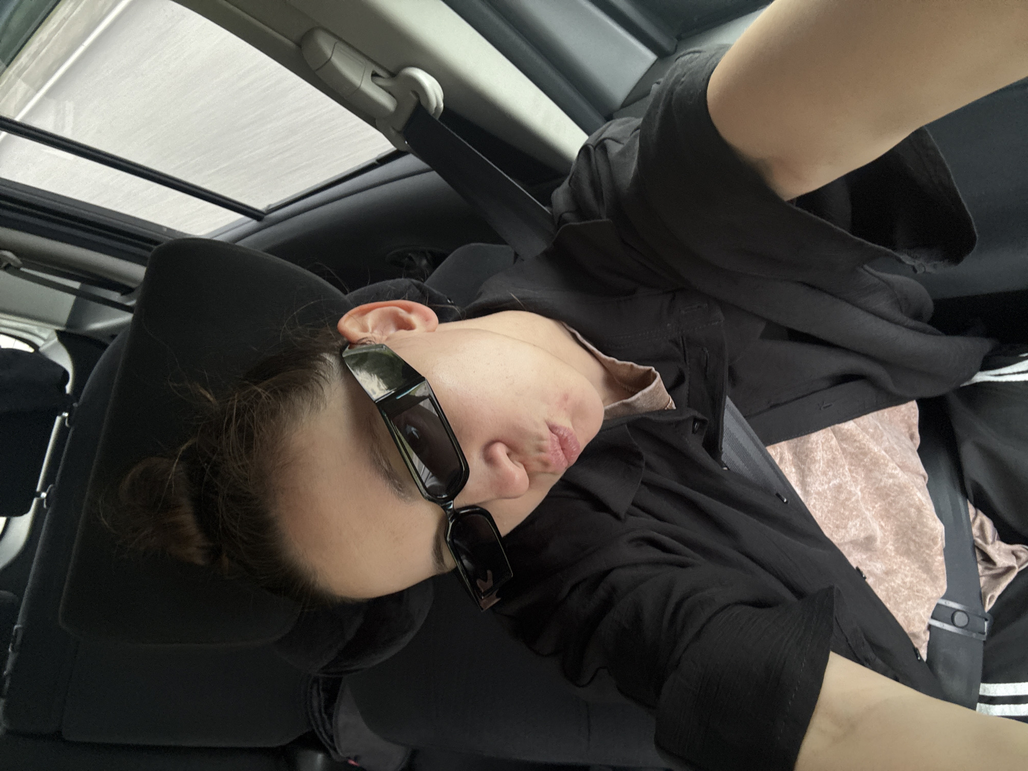 A person is inside a car, leaning against the headrest. The person is wearing sunglasses and a black shirt, and a seatbelt is visible. The car has a sunroof. The location is not possible to determine.
