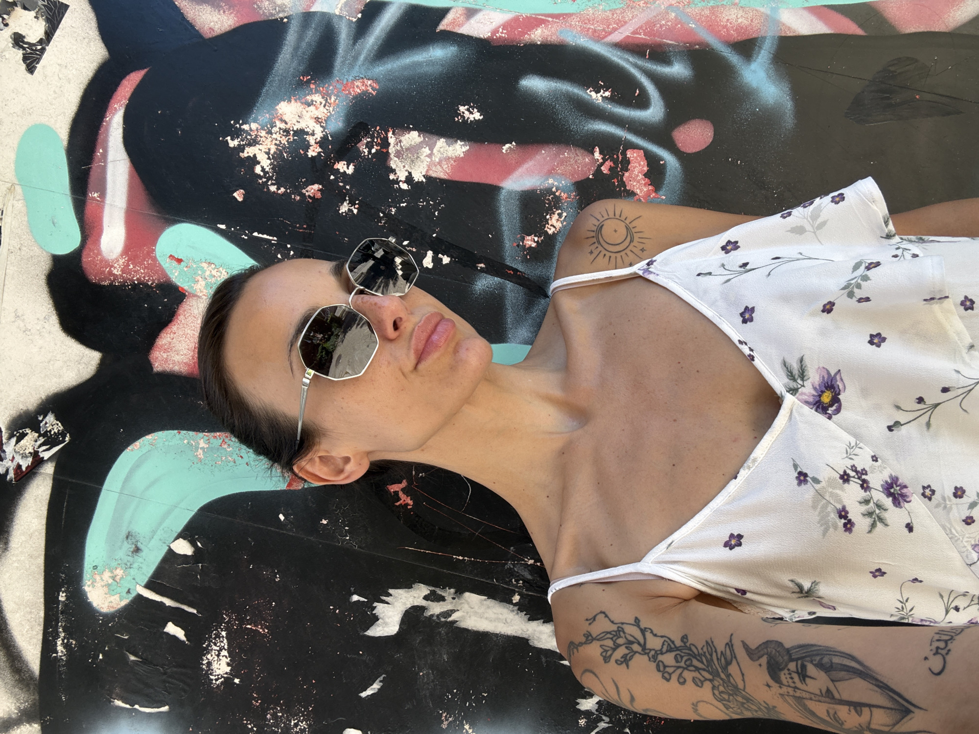 A woman with dark hair pulled back, wearing large, silver-rimmed sunglasses and a white floral-print spaghetti strap dress, poses in front of a graffiti-covered wall. The graffiti features large abstract shapes in black, teal, and red. The woman has tattoos on her left arm and shoulder, including a sun and moon design on her shoulder. The scene appears to be outdoors, likely in an urban setting. The location is identified as Pleven, Bulgaria, based on external metadata.
