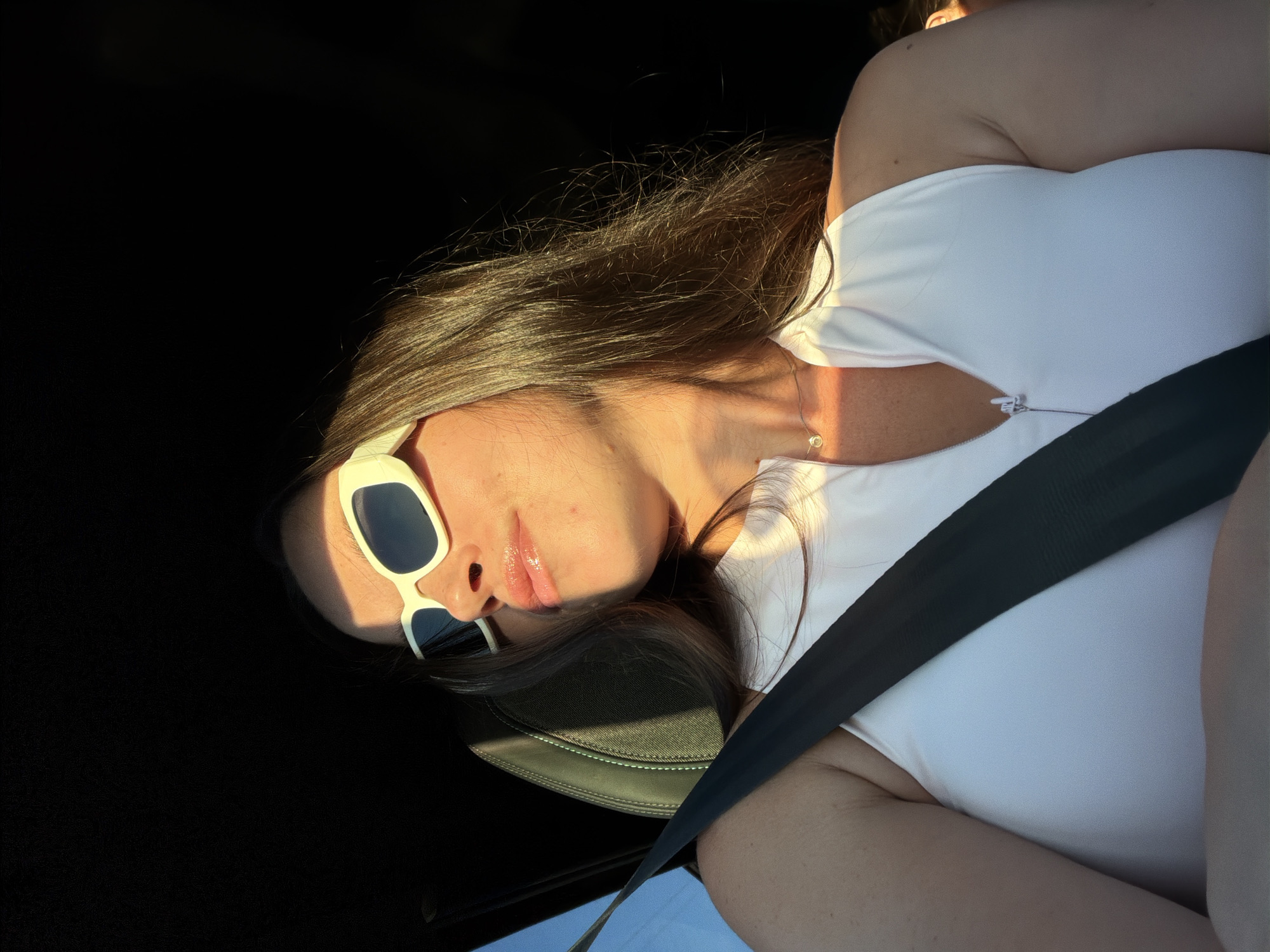 A medium shot captures a woman from the chest up, seated in a car. She is wearing white sunglasses with thick frames, a white zip-up halter top, and a delicate silver necklace with a small circular pendant. Her long, brown hair falls over her shoulders. The harsh sunlight casts strong shadows across her face and upper body. A black seatbelt is visible across her chest. The background is dark, suggesting the interior of a car. The image was taken in Vinyols i els Arcs, Spain.