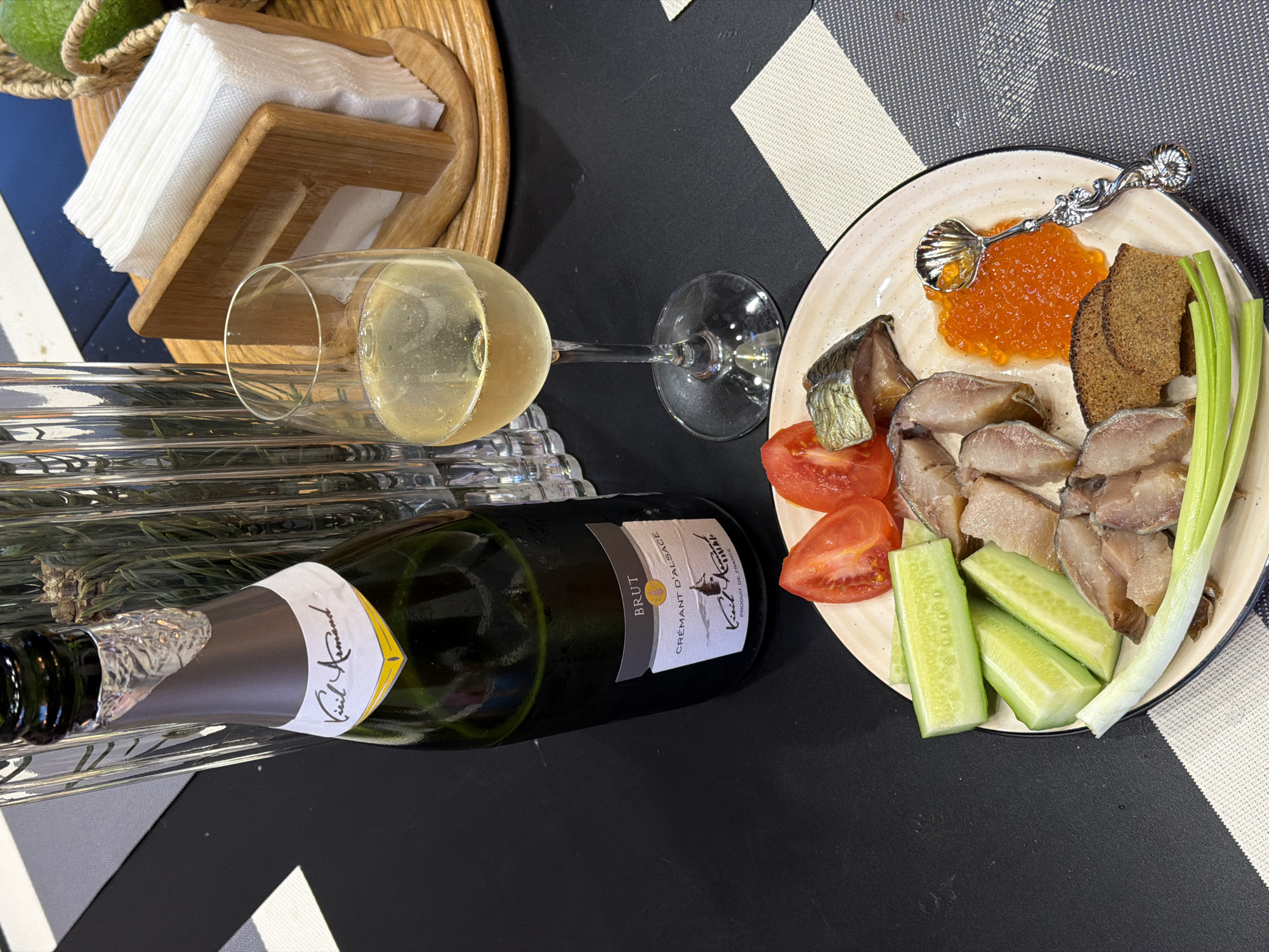 The image shows a table setting with food and drink. A bottle of "Kiel Armand Cremant d'Alsace Brut" lies horizontally near a crystal glass filled with a light-colored sparkling wine. A plate holds a variety of foods: what appears to be sliced fish, red caviar with a decorative silver spoon, a piece of dark bread, scallions, slices of tomato, and cucumber sticks. A wooden napkin holder containing white napkins sits on the upper left. The table surface has a black and white patterned covering. The location is unspecified but there is no forensic indication that it is set in the city of Ryazan, Russia.