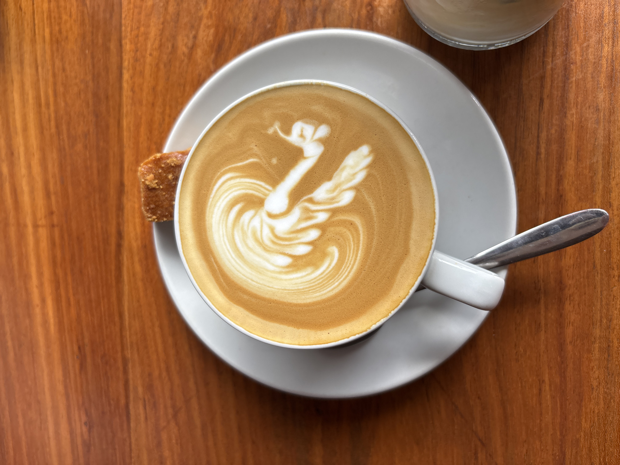 A top-down shot shows a cup of coffee with latte art resembling a swan on a saucer with a small biscuit or pastry, and a spoon on a wooden table. Another glass is partially visible in the upper right corner. The location, Kuta Selatan, Indonesia, does not influence the visual elements significantly. The main subjects are the coffee and its presentation, the accompanying pastry, and the tableware. The environment is a simple wooden table. There are no people visible in the image. No actions or interactions are directly observed, as the scene is static.
