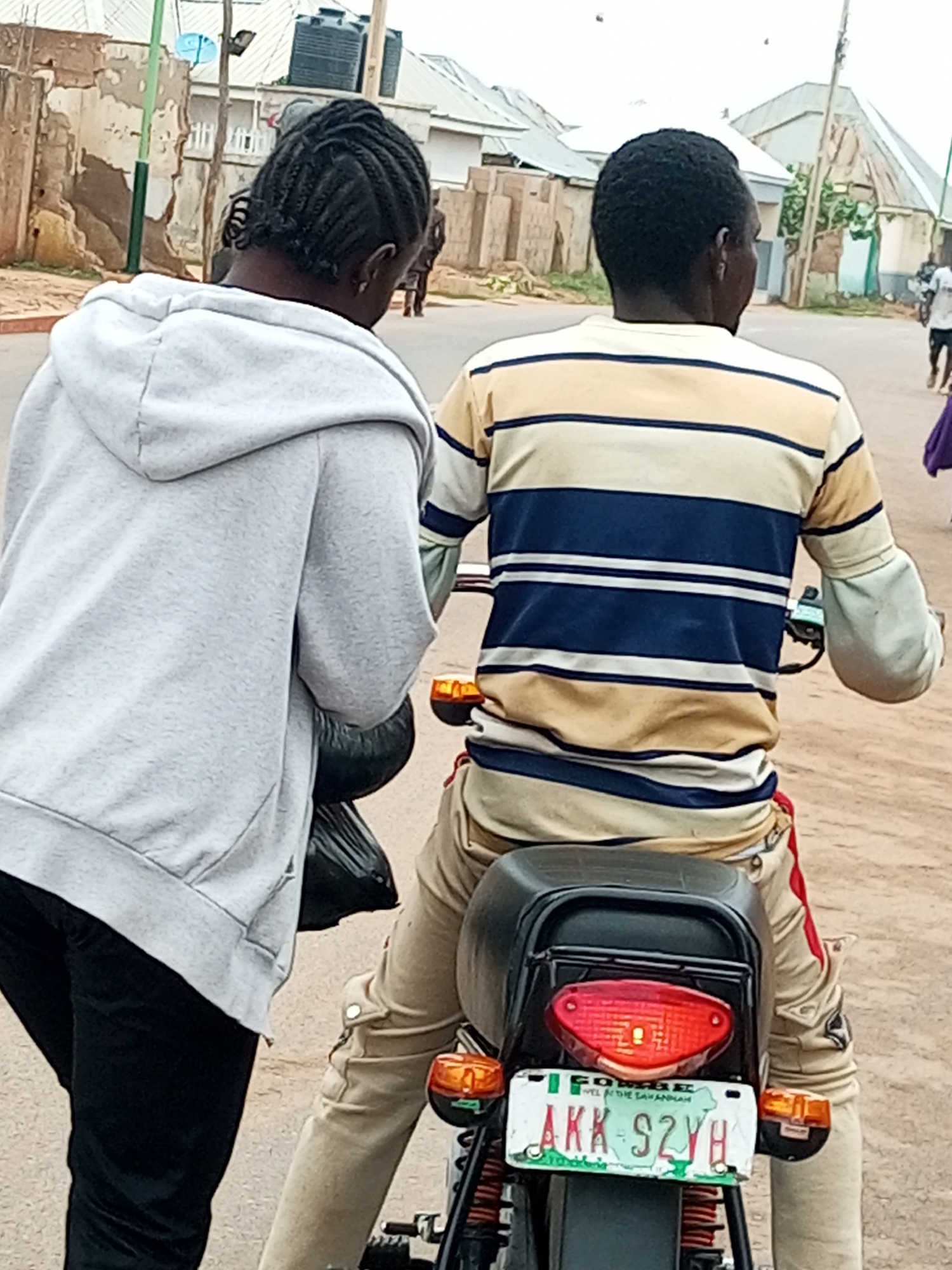 In Tempure, Nigeria, two individuals are seen from behind. A person on the left wears a light grey hooded sweatshirt and black pants, carrying a black plastic bag. To their right, a person is seated on a motorcycle, wearing a striped long-sleeved shirt in yellow, blue, and white, and light brown pants with red accents. The motorcycle has a visible license plate that reads "AKK S2YH". The scene appears to be on a dusty street with buildings and structures in the background.