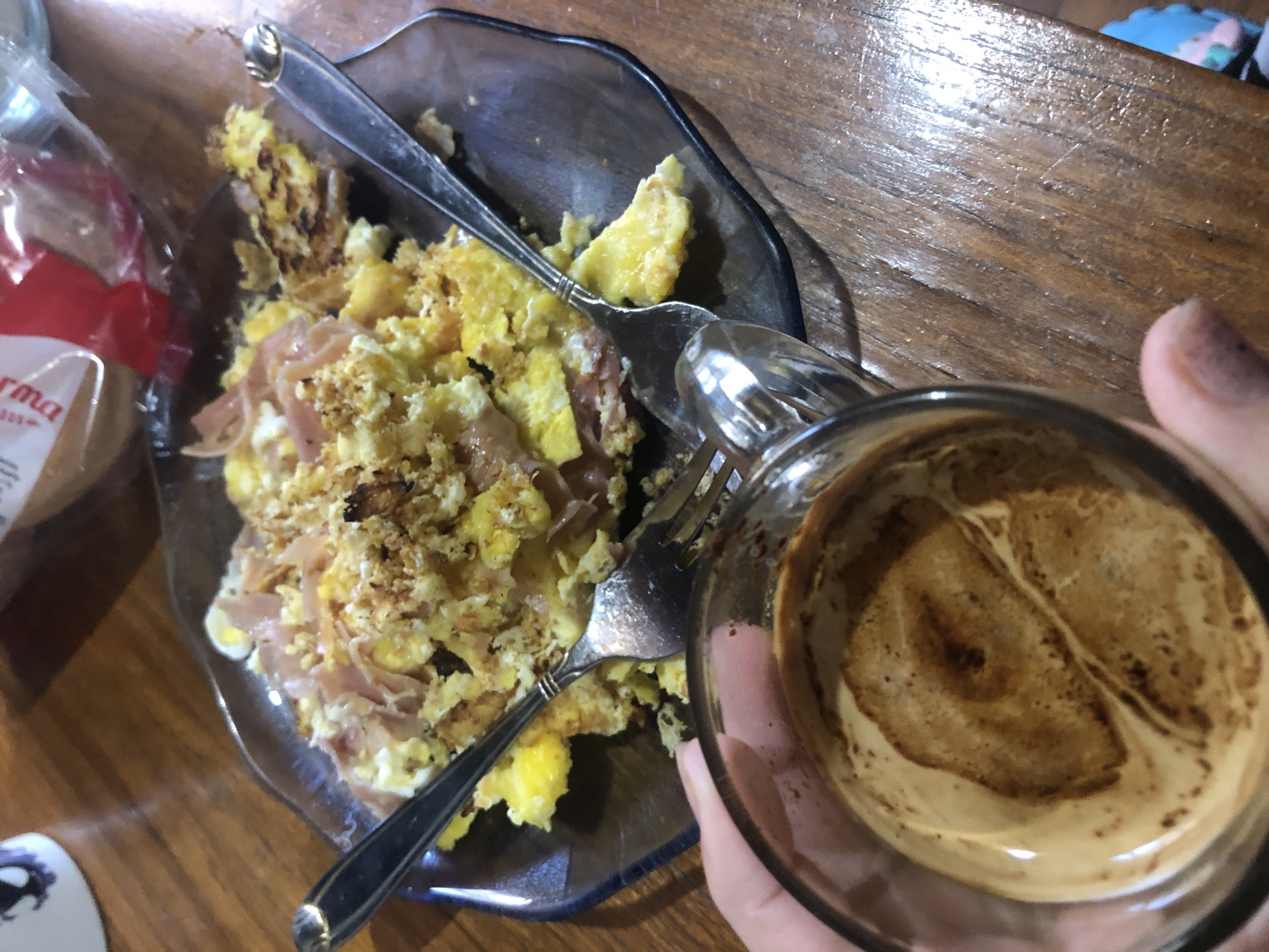 A plate of scrambled eggs with ham and breadcrumbs is placed on a wooden table in an indoor environment. Two silverware pieces are inside the plate of food. A clear glass of coffee with cream is held to the lower-right of the plate of food. A package of sliced bread is to the left of the plate. The location is implied to be in Buenos Aires, Argentina. There are no visible people. No specific actions are depicted.