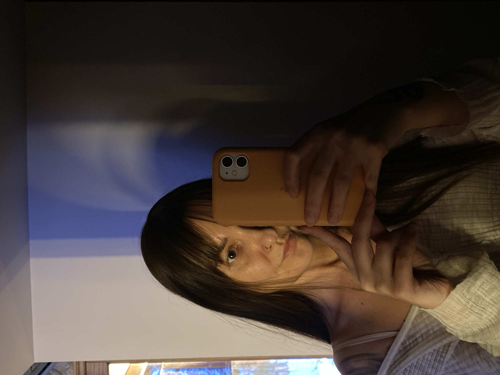 A woman with dark hair and bangs is taking a selfie using an orange phone. She is holding the phone in her left hand, partially obscuring her face. She has a nose ring. She is wearing a light-colored, textured top and a white spaghetti strap. A glimpse of a tattoo is visible on her upper chest. The background is divided into a light area on the left and a darker area on the top. A multi-colored light source can be seen at the bottom of the image. The location is not discernible from the image. No evidence points to Votkinsk, Russia.