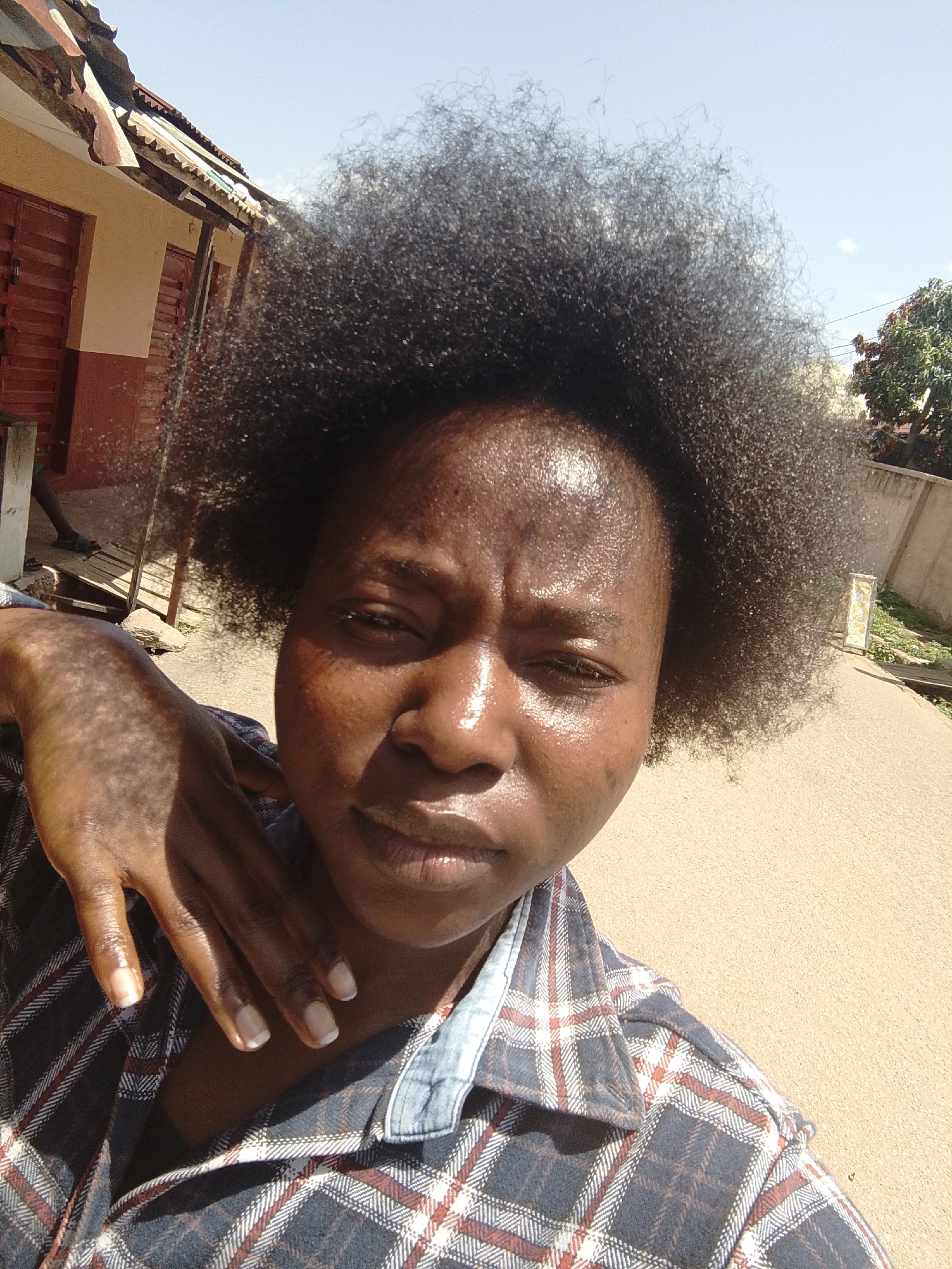 The image shows a close-up selfie of a young Black woman with very large, dark, curly hair. She is wearing a plaid shirt with blue, white, and red patterns. Her skin is dark and appears shiny, possibly from sweat or product. She has a hand raised to her face, with her fingers splayed. Her fingernails are neatly manicured with a light polish. The background suggests an outdoor setting, with a building to the left and a dirt or gravel path extending into the distance on the right. The lighting indicates it is daytime, likely sunny. The image was captured in Jos, Nigeria.