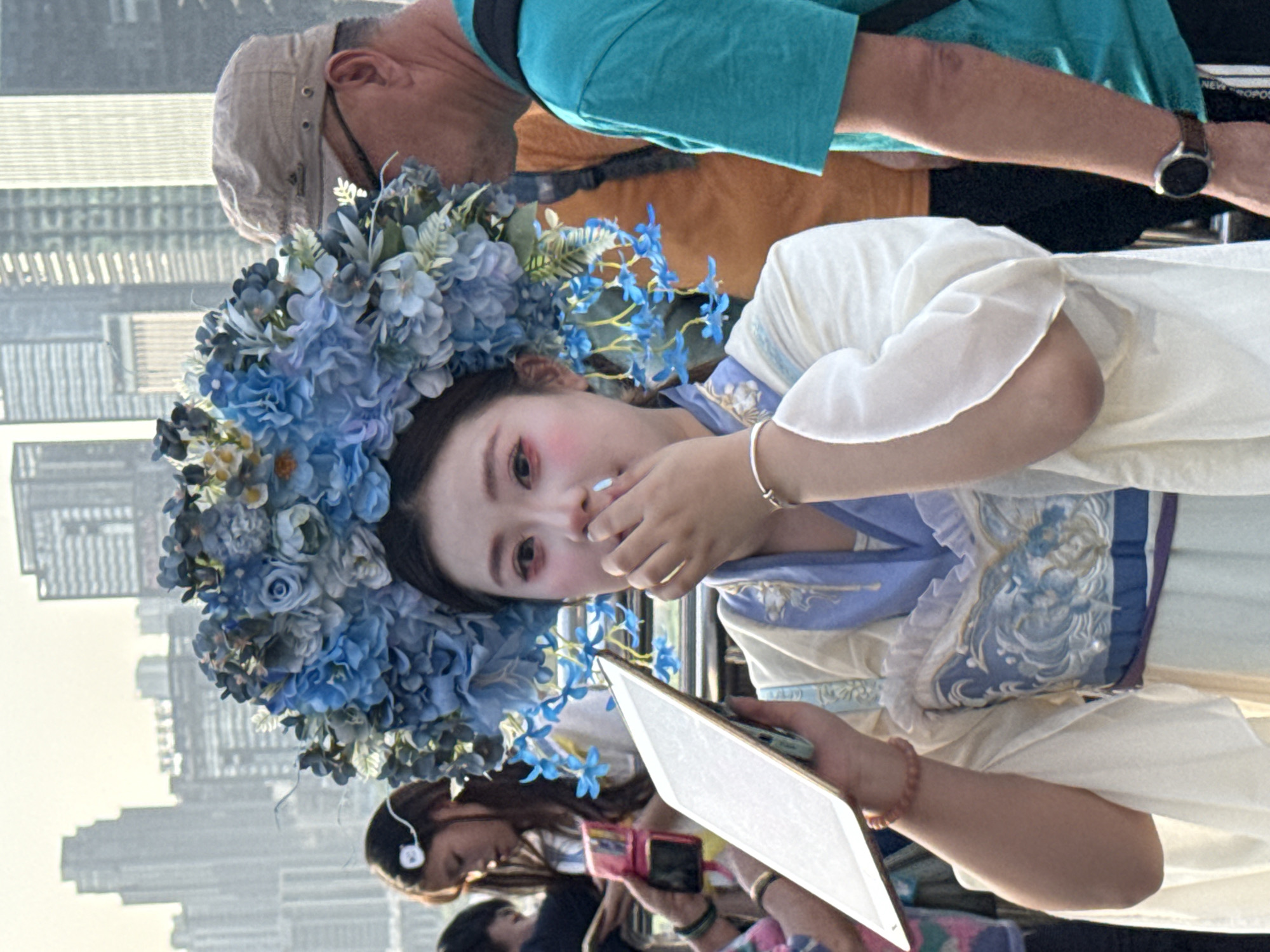An outdoor, portrait-oriented photograph captures a young woman in the foreground, wearing a large, elaborate headpiece made of blue and light-colored artificial flowers. She is dressed in a light-colored, possibly traditional Chinese garment, resembling a Hanfu, with blue and purple embroidered details on the bodice. Her left hand is raised near her mouth, and her right hand holds a white tablet or smartphone. She looks directly at the camera with a neutral expression, enhanced by subtle makeup and blush. A silver bracelet is visible on her left wrist, and a beaded one on her right.
In the mid-ground, an older man is partially visible, wearing a light-colored cap, a teal t-shirt over a brown shirt, and a watch on his left wrist. He appears to be facing away from the camera. The background is a dense, blurred urban landscape featuring numerous tall buildings under what appears to be soft, late afternoon or early evening light. Other blurred figures, including a woman with headphones and another holding a pink phone, are discernible further back. The visible text "NEW PROPOS" is partially seen on the older man's black strap. The scene suggests a cultural event or a photoshoot taking place in a bustling cityscape, characteristic of Yuzhong District, China.