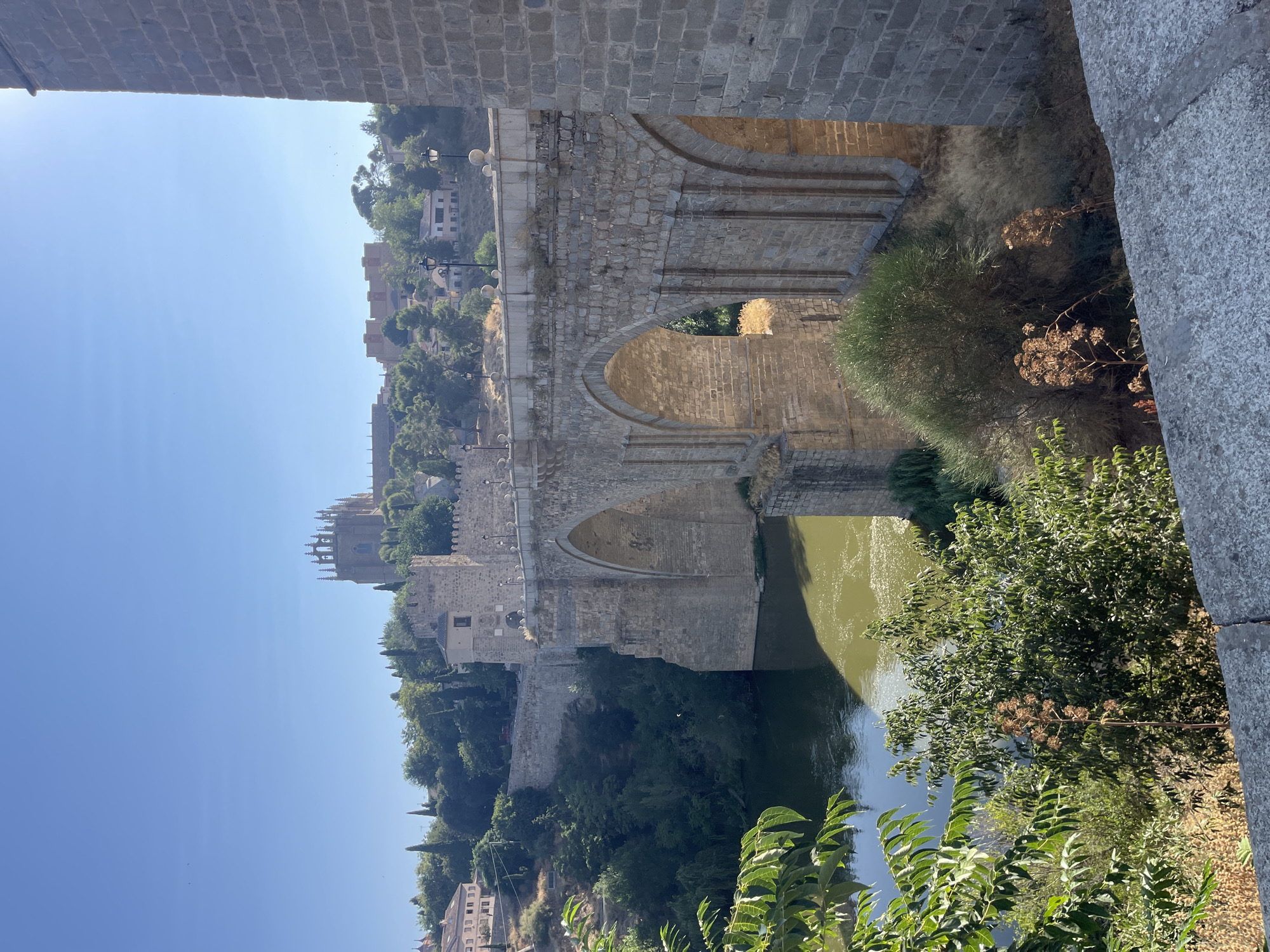 The image shows the San Martín Bridge in Toledo, Spain, a medieval stone bridge that spans the Tagus River. The bridge is characterized by its pointed arches and robust stone construction. In the background, the city of Toledo is visible, with its historic buildings and a prominent cathedral with Gothic spires silhouetted against a clear blue sky. The river below the bridge has a greenish hue, reflecting the sunlight. Lush green trees and vegetation line the riverbanks and the slopes of the city, adding to the scenic beauty of the location. The foreground features a portion of a stone wall and dry vegetation, suggesting a warm, possibly arid climate. The overall scene depicts a historic landmark in a well-preserved urban setting, bathed in bright daylight.