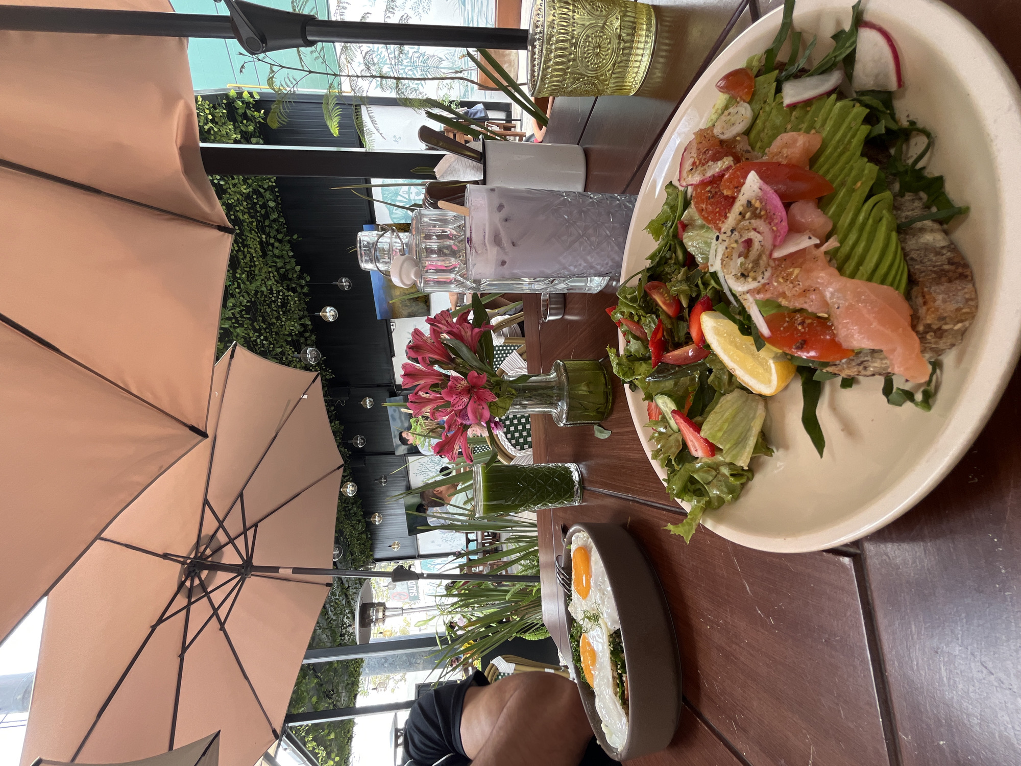The image shows a table at an outdoor restaurant, likely in San Bernardino Tlaxcalancingo, Mexico, based on the presence of bougainvillea and other regional flora. A large beige patio umbrella shades the scene. On the table are a plate of salad with salmon and avocado, a plate of fried eggs, a tall green smoothie, a vase of pink alstroemeria flowers, a water carafe, a glass of purple liquid, and a container with utensils. People are visible in the background, seated at other tables. The table is dark wood. The overall setting is bright and sunny. A person's arm is visible in the lower left corner of the image.