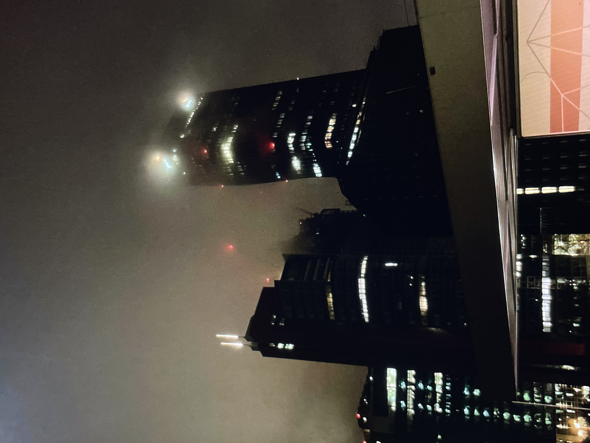 The image shows a night view of city buildings shrouded in fog. The buildings are mostly dark with illuminated windows creating vertical streaks of light. Some buildings appear to be skyscrapers, recognizable by their height and rectangular shape. The fog obscures the upper parts of these buildings, creating a sense of mystery. There are red lights visible on the tops of some buildings, which might be safety or aviation lights. The sky is a uniform gray due to the heavy fog. The perspective suggests the photo was taken from a high vantage point, possibly from another building.