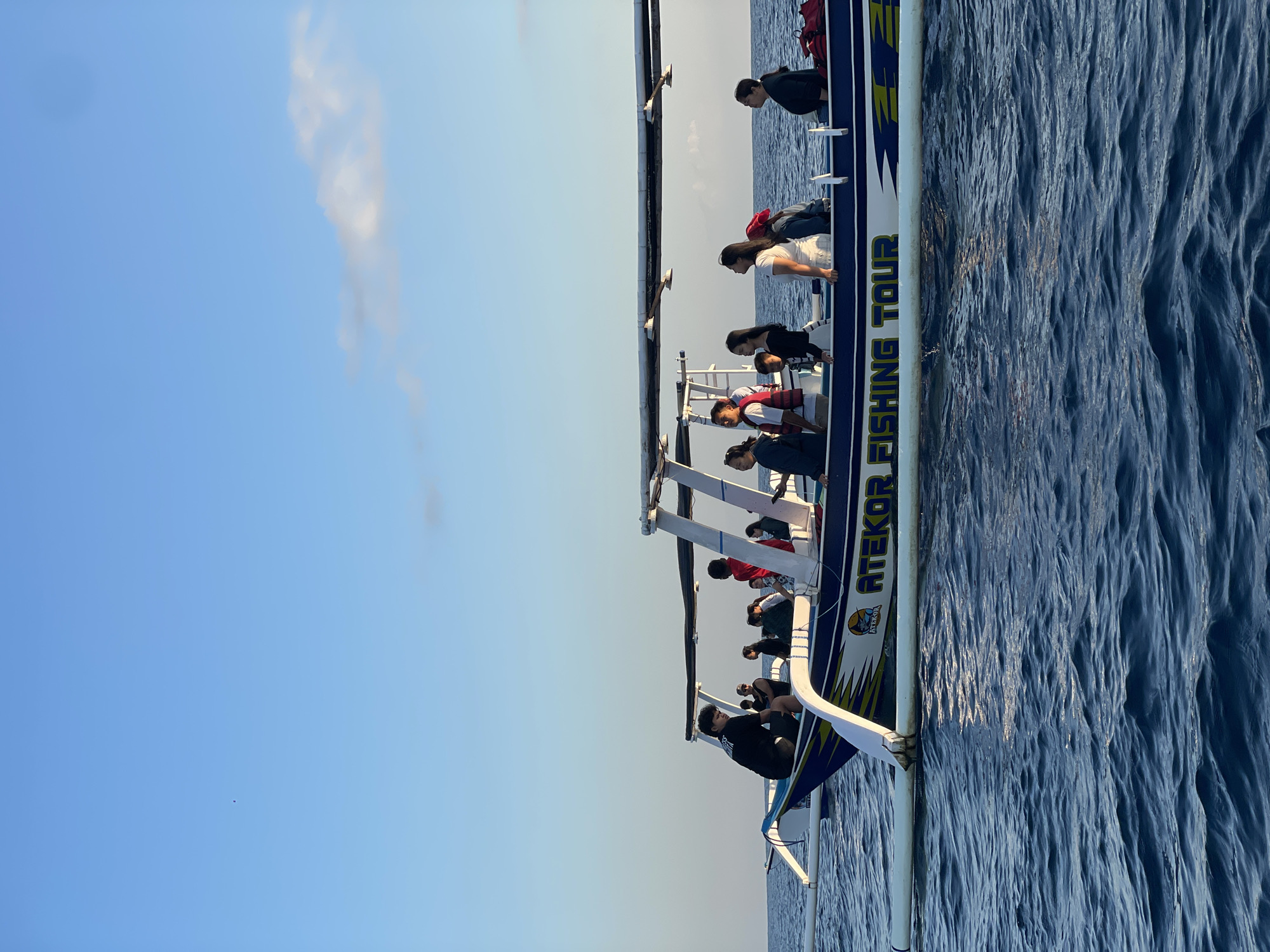 In an outdoor, medium-shot, eye-level perspective, a group of people are aboard a white and blue fishing boat named "ATEKOR FISHING TOUR" as it sails on the ocean under a clear, light blue sky. The boat, characterized by its outriggers, features a canopy supported by white poles. Several individuals are visible on the boat, some standing and others seated, with their faces turned in various directions. The water is a deep blue, with small ripples creating a textured surface. The sky is mostly clear, with a faint cloud formation present near the horizon on the right side of the image. The scene is set during daylight, likely late afternoon or early morning given the soft lighting and the quality of the sky. The location is identified as Unknown, Indonesia.