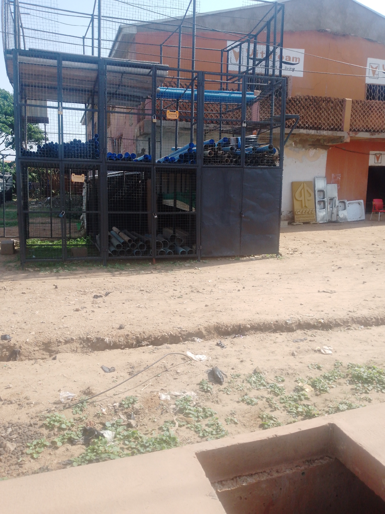 The image shows an outdoor storage cage filled with pipes, located in Sabon Tasha, Nigeria. The cage appears to be constructed of metal and contains blue and grey pipes. The storage cage is located on a dirt surface, with some vegetation nearby. Behind the cage, a building with an orange facade is visible. A sign with the words "Vitam First Living" is on the building. Sinks are stacked near the building behind the cage. A red chair is visible in front of an open door leading into the building. A drainage ditch is present in the foreground.
