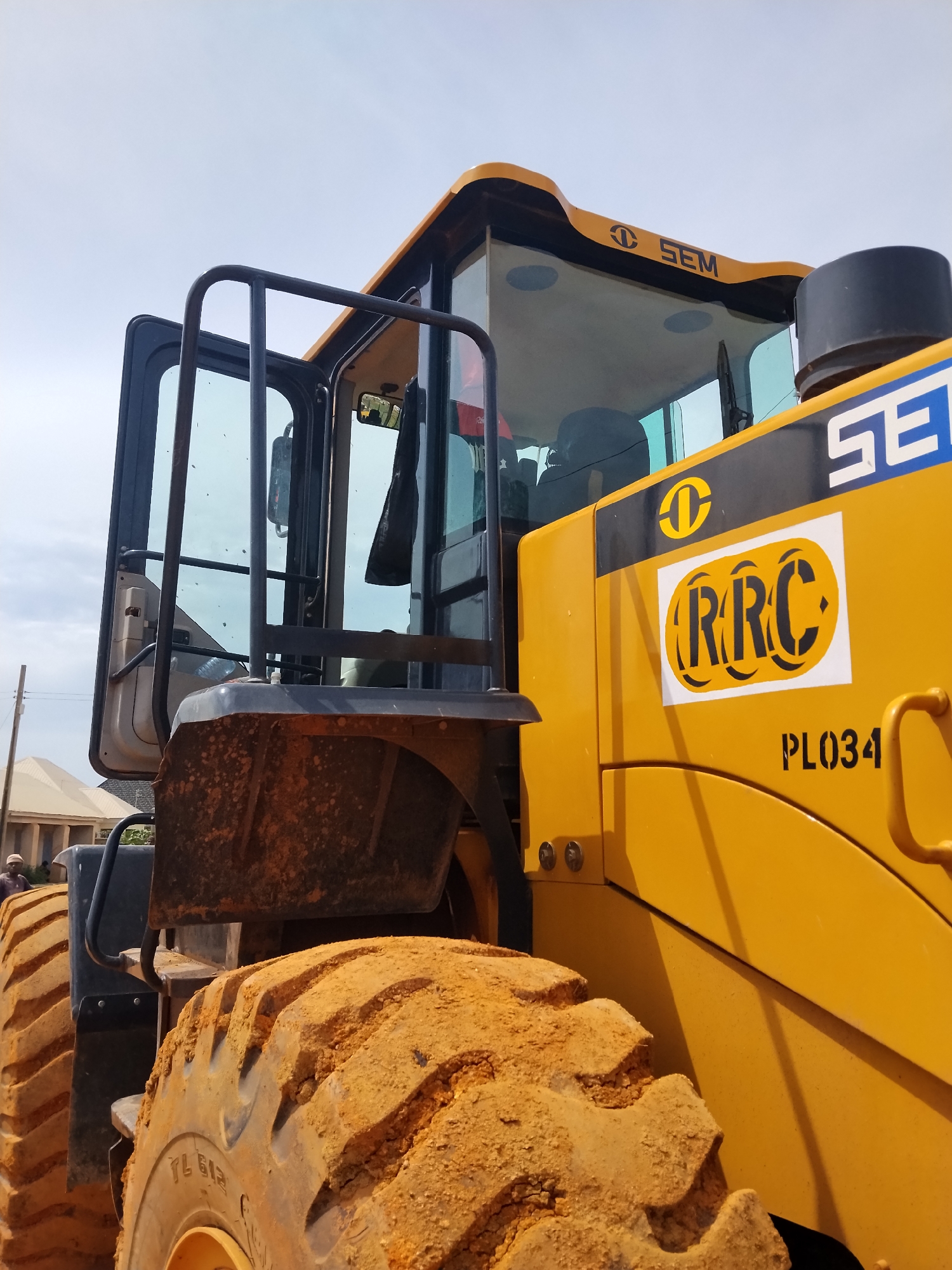 A close-up, low-angle, side view captures a yellow SEM brand front loader vehicle. The vehicle is positioned in a city, likely Laminga, Nigeria, with buildings visible in the background. The foreground is dominated by the vehicle's large, orange, mud-caked tire and a portion of its yellow bodywork, which features black stenciled text reading "PL034". Above the tire, the cab of the front loader is visible, with its door ajar, revealing part of the interior. The cab also displays the SEM logo and branding. The sky above is a light blue with some clouds. The overall scene suggests an operational or recently used construction or heavy machinery site.