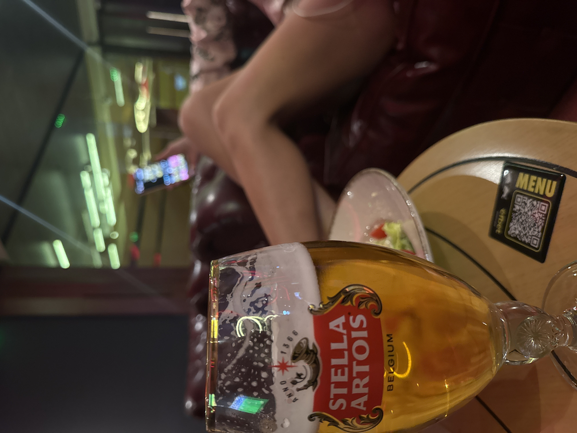 The image depicts an interior scene, possibly a bar or restaurant, featuring a partially consumed glass of Stella Artois beer in the foreground. Behind the beer, there's a small plate with food, likely a salad. A light-colored wooden table is visible, with a menu featuring a QR code resting on it. In the blurred background, a person is seated on a red leather-like couch, possibly using a phone. Bright, out-of-focus lights suggest an urban setting, potentially in Sofia, Bulgaria. There is no concrete evidence in the image that would specify it is in Sofia, Bulgaria.
