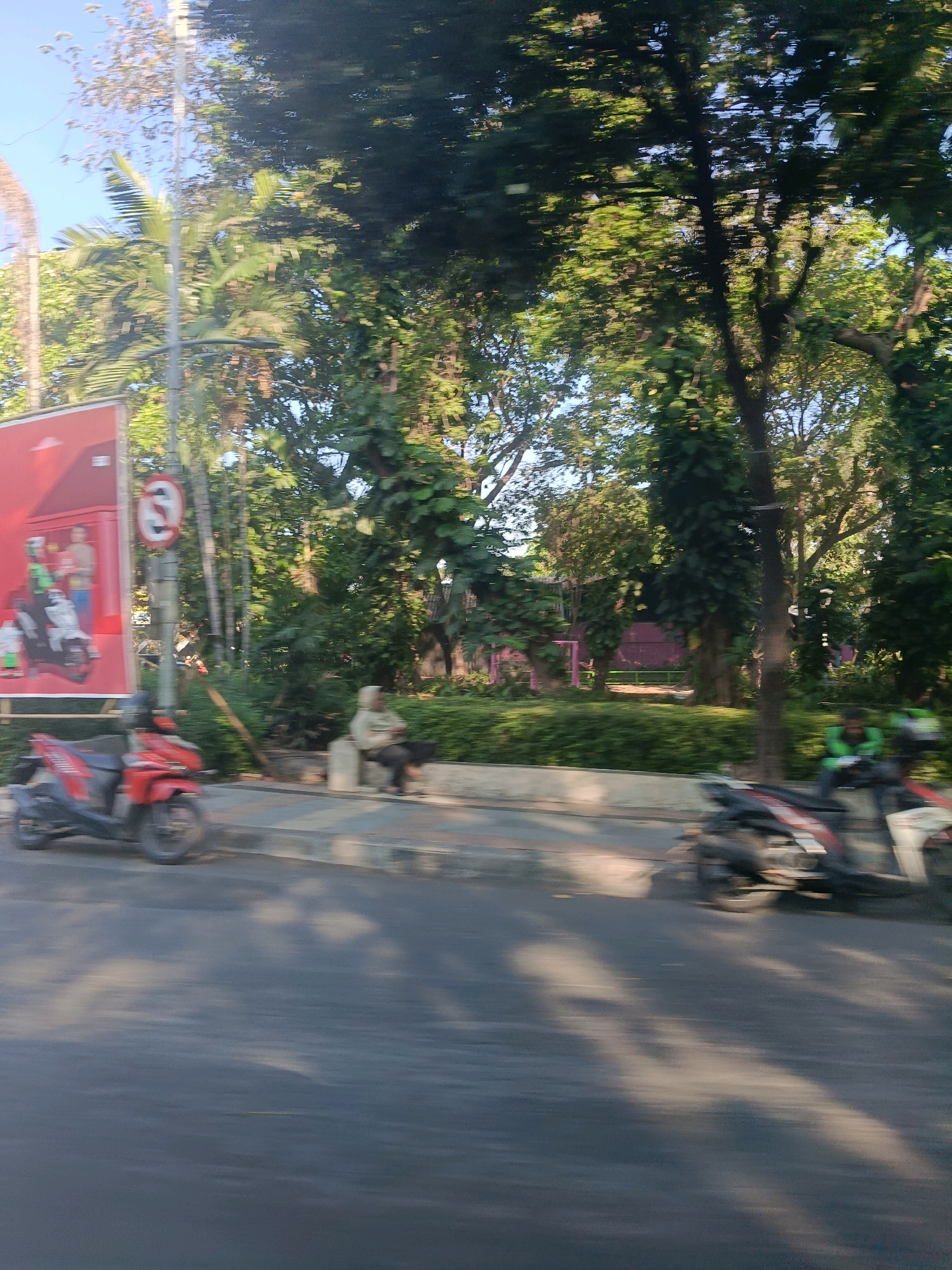 A slightly blurred, eye-level, outdoor daytime shot shows a street scene in Surabaya, Indonesia. The foreground is dominated by a dark gray asphalt road with dappled sunlight and shadows from trees. To the left, a bright red scooter is parked on a sidewalk, which is paved with patterned gray and light gray tiles. Behind the scooter, a large, vertical billboard features a red background with images of people and a motorcycle. A traffic sign with a circular red border and a diagonal red line over a black "E" symbol is mounted on a pole to the right of the billboard. Further down the sidewalk, a person in light-colored clothing is seated on a light gray bench.
Beyond the sidewalk, lush green trees and foliage form a dense canopy, with some smaller trees and bushes visible. In the background, parts of buildings or structures with purple and pink accents are partially visible through the trees.
On the right side of the road, another scooter is partially visible, with a person wearing a green jacket and white pants seated on it. The overall impression is a busy urban environment with ample greenery.