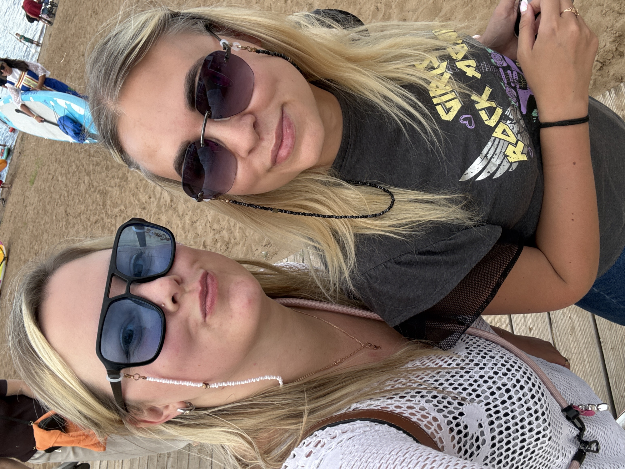 Two women are taking a selfie at a beach in Chaikovsky, Russia. The woman in the foreground is wearing large blue-tinted sunglasses and a white crocheted top. She has blonde hair and is wearing a delicate necklace. The woman behind her is wearing darker sunglasses and a grey t-shirt with a graphic print. She also has blonde hair and is wearing a beaded chain around her neck. The background shows a sandy beach with people in the distance, some of whom appear to be on paddleboards or kayaks. The overall mood is casual and relaxed, suggesting a sunny day at the beach. The graphic on the t-shirt of the woman in the background has text that appears to say "BAD AXE" in yellow lettering with a stylized winged design.