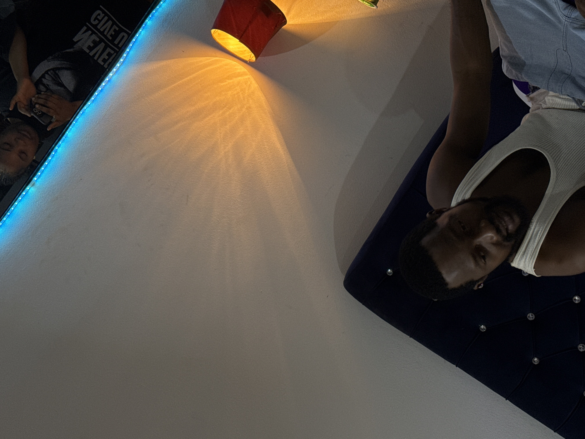 A low-angle shot reveals a man lying on a dark blue velvet bed or headboard adorned with decorative buttons, his face and upper body visible. He wears a white tank top and appears to be looking at the camera. Above him, on the wall, a small red lamp emits a warm glow, casting a pattern of light onto the pale wall. To the left, a mirror or framed picture hangs, reflecting a person using a phone. The room features a blue LED strip light, accentuating the edge of the mirror or frame. The environment seems to be an interior space, possibly a bedroom. The location is unknown, and there is no evidence to suggest this image was taken in Abuja, Nigeria.