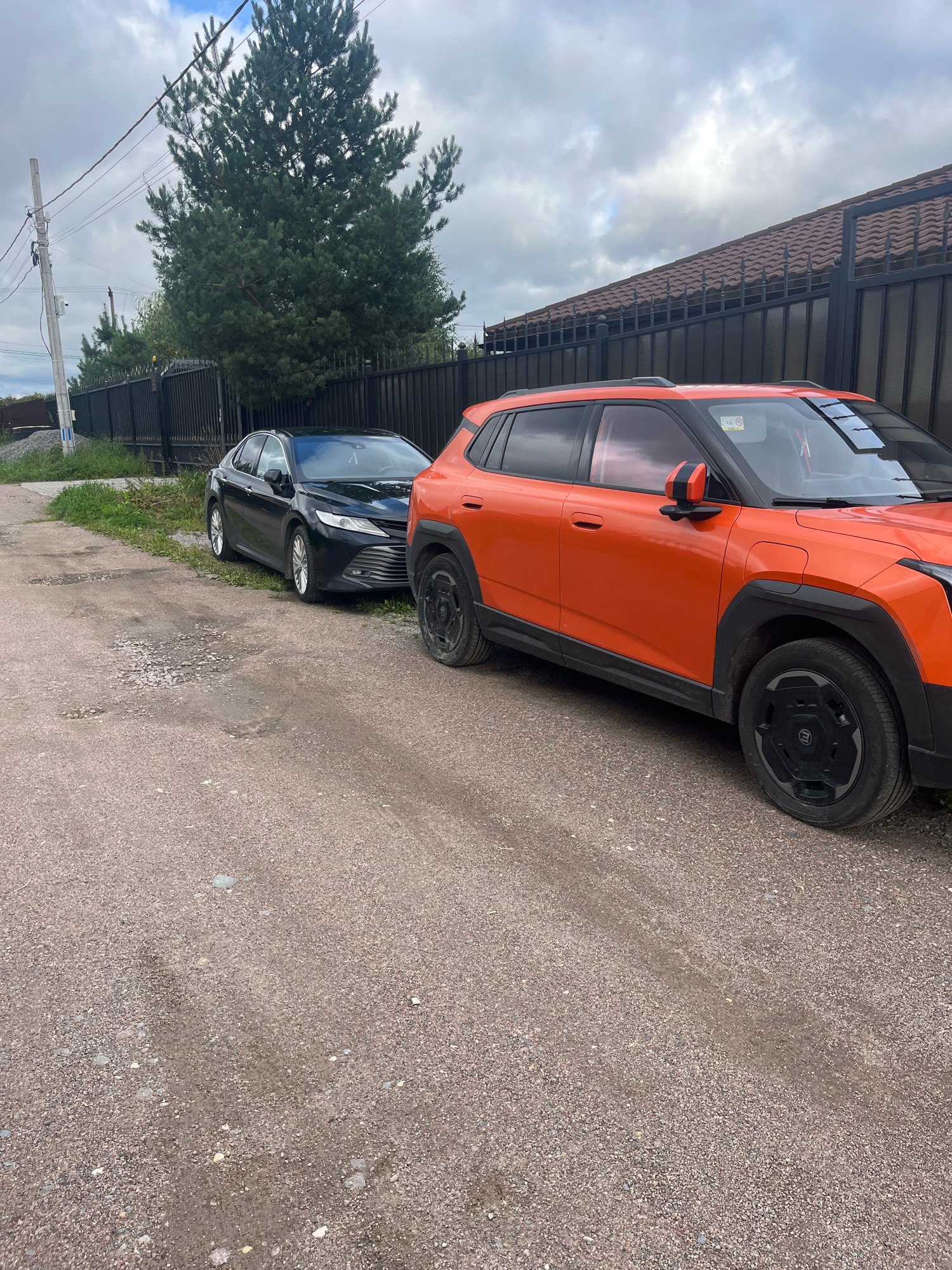 Two parked cars, a black sedan and an orange SUV, are situated on a gravel road next to a black metal fence with a tiled roof on top. A large evergreen tree is visible behind the fence and to the left of the cars. The sky is overcast with clouds. The scene appears to be in a residential area.