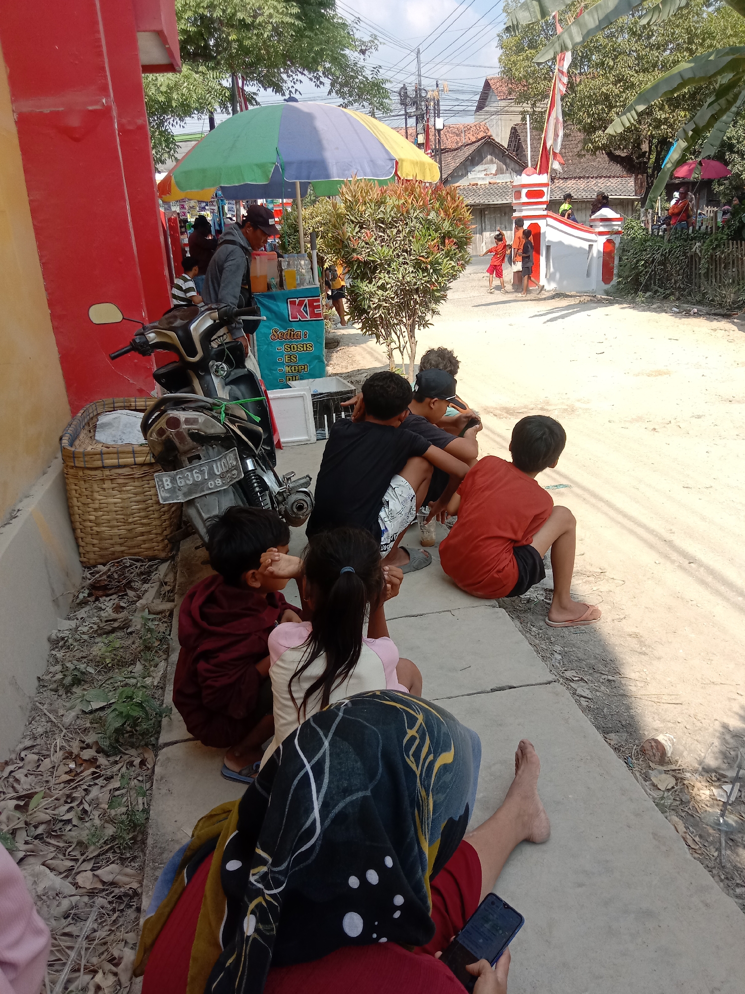 In Kunden, Indonesia, on a sunny day, a group of children is gathered by a roadside stall. A man in a gray hoodie and a black cap stands behind the stall, which is adorned with an umbrella and displays various drinks. A motorcycle is parked nearby. The children, dressed in casual clothing, are sitting on the concrete sidewalk, some facing the street, others looking towards the stall. In the background, a white and red gate stands with an Indonesian flag fluttering from a pole. The scene is a slice of everyday life in the city, possibly during a local event or simply a typical afternoon.