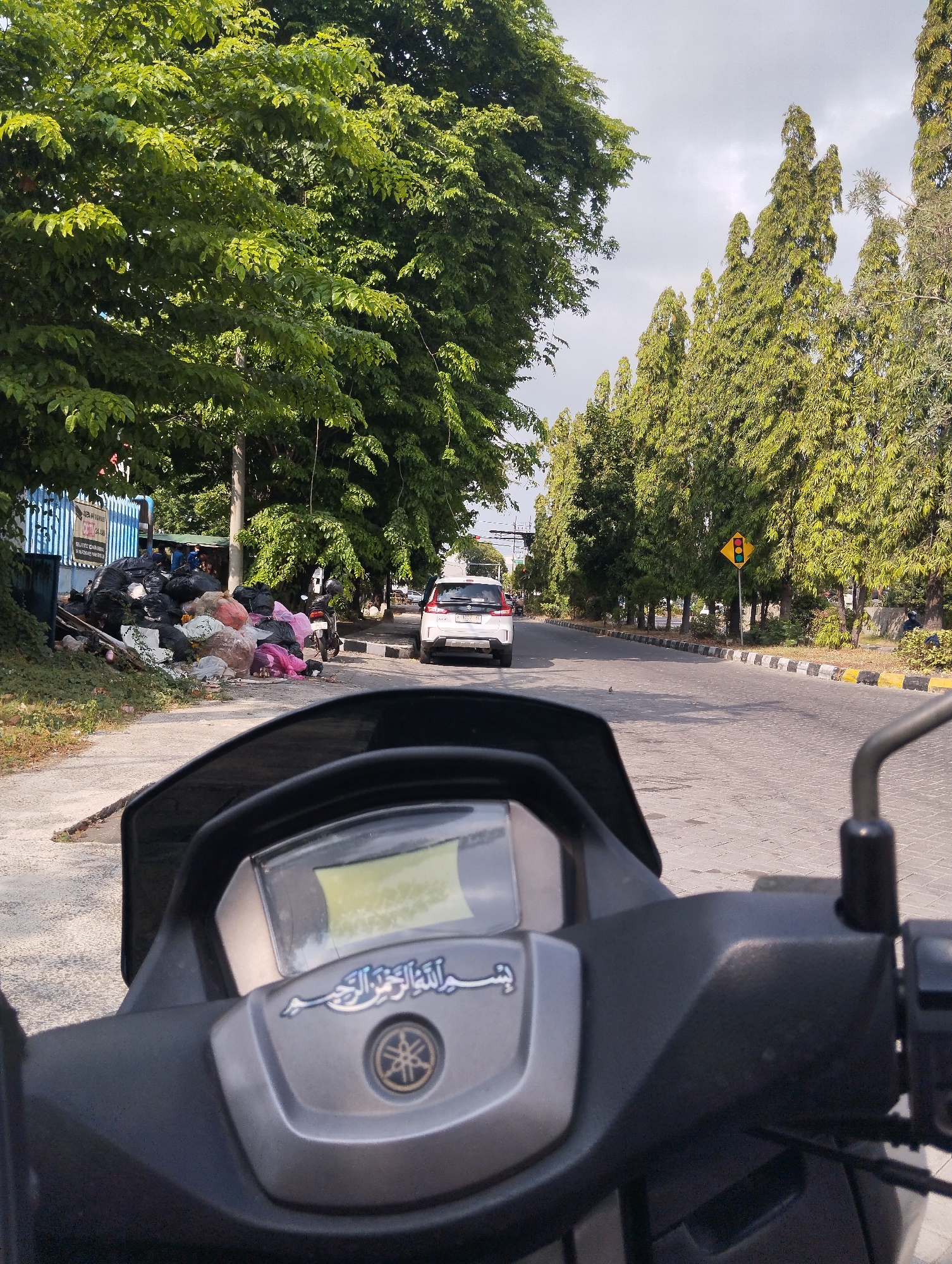 The image appears to be taken from the perspective of someone riding a motorcycle, looking forward down a paved road lined with trees. On the left side of the road, there is a large pile of garbage bags, some pink and some black, next to a blue building. Further down the road, there are several tall, thin trees with green foliage. A white car is visible in the distance, and a traffic light is on a pole to the right of the road. The foreground of the image shows the handlebars and instrument panel of the motorcycle. Based on the presence of the Yamaha logo on the motorcycle's panel and the general context, the location is likely a street in Surabaya, Indonesia.