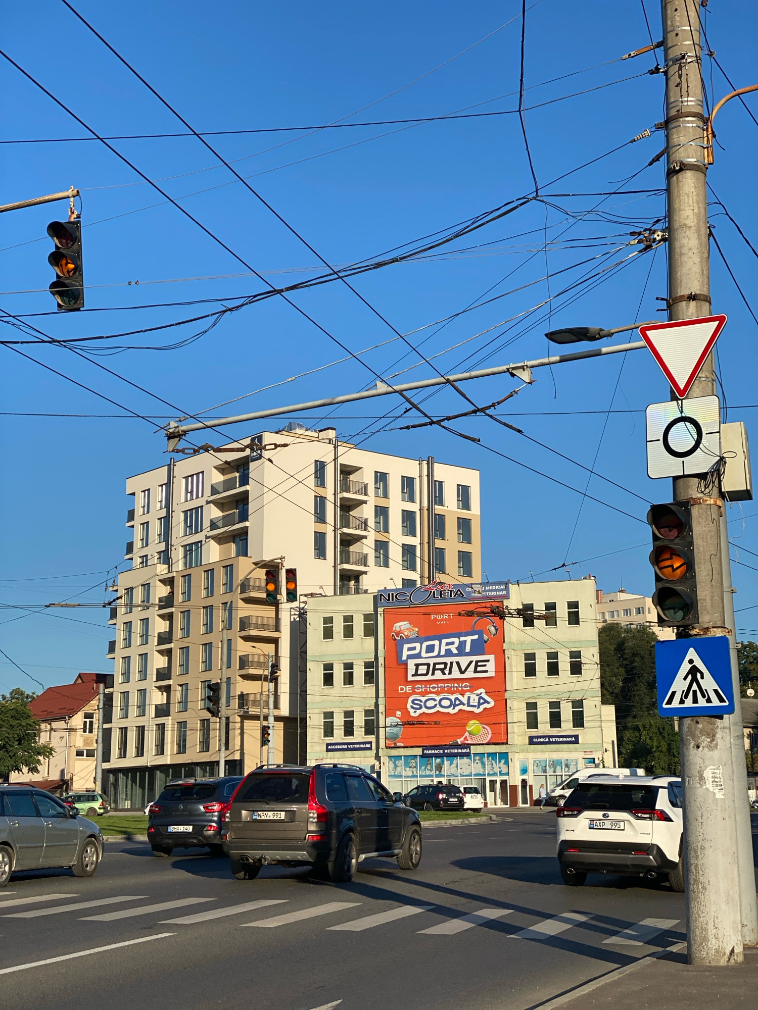 The image depicts a street scene in Chișinău, Moldova, on a clear day with a bright blue sky. The primary focus is an intersection with a prominent traffic light pole on the right side. This pole displays multiple traffic signals, one of which is illuminated yellow. Overhead, a complex network of electrical wires crisscrosses the sky, characteristic of urban infrastructure.
A multi-story modern apartment building, characterized by its beige and white facade and numerous balconies, dominates the background. Adjacent to it are other buildings, including one with a large orange billboard advertising "PORT DRIVE DE SHOPPING ȘCOALA," and another with signs for "CLINICA VETERINARĂ" and "FARMACIE VETERINARĂ" displaying images of animals.
In the foreground, several cars are visible at the intersection, including a silver hatchback on the left, a dark-colored SUV in the center, and a white SUV on the right. A pedestrian crossing is marked with white stripes on the asphalt. Traffic signs are also present, including a yield sign and a pedestrian crossing sign. The overall impression is a busy urban intersection in daylight.