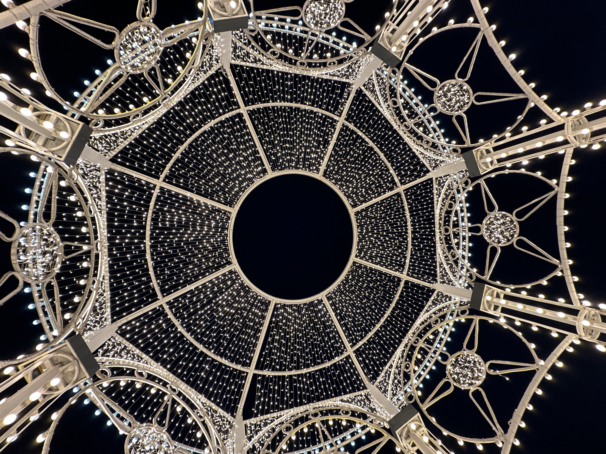 An upward-looking photograph shows a white metal light structure against a black sky. The structure is circular with an open center, resembling a dome or gazebo. It is illuminated with numerous small, white lights strung in lines. At the perimeter, there are larger decorative elements, also lit with small lights. The overall scene appears to be a public space, possibly a holiday or festival display. The location is unknown, and the image does not contain any people.