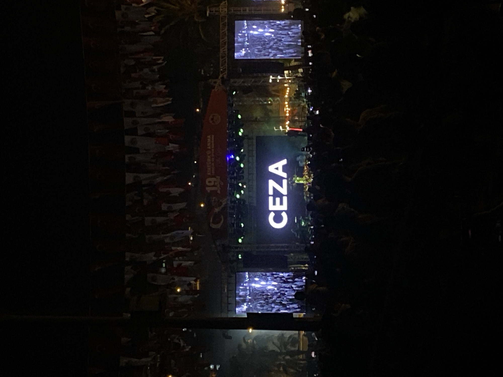 The image captures a nighttime scene at what appears to be an outdoor concert or event. The focus is on a stage featuring the illuminated name "CEZA" in large, bold letters. Above and below, screens display visual content, possibly related to the performance. The audience is visible on both sides of the stage, with many individuals holding up their phones, presumably to record or photograph the event. The backdrop includes a vertical banner bearing the words "ATATÜRK'Ü ANMA" along with the number "19," indicating a commemoration. Further to the left, a multitude of Turkish flags are arrayed in the night. The setting is likely Alanya, Turkey, based on the presence of Turkish flags and Atatürk commemoration.