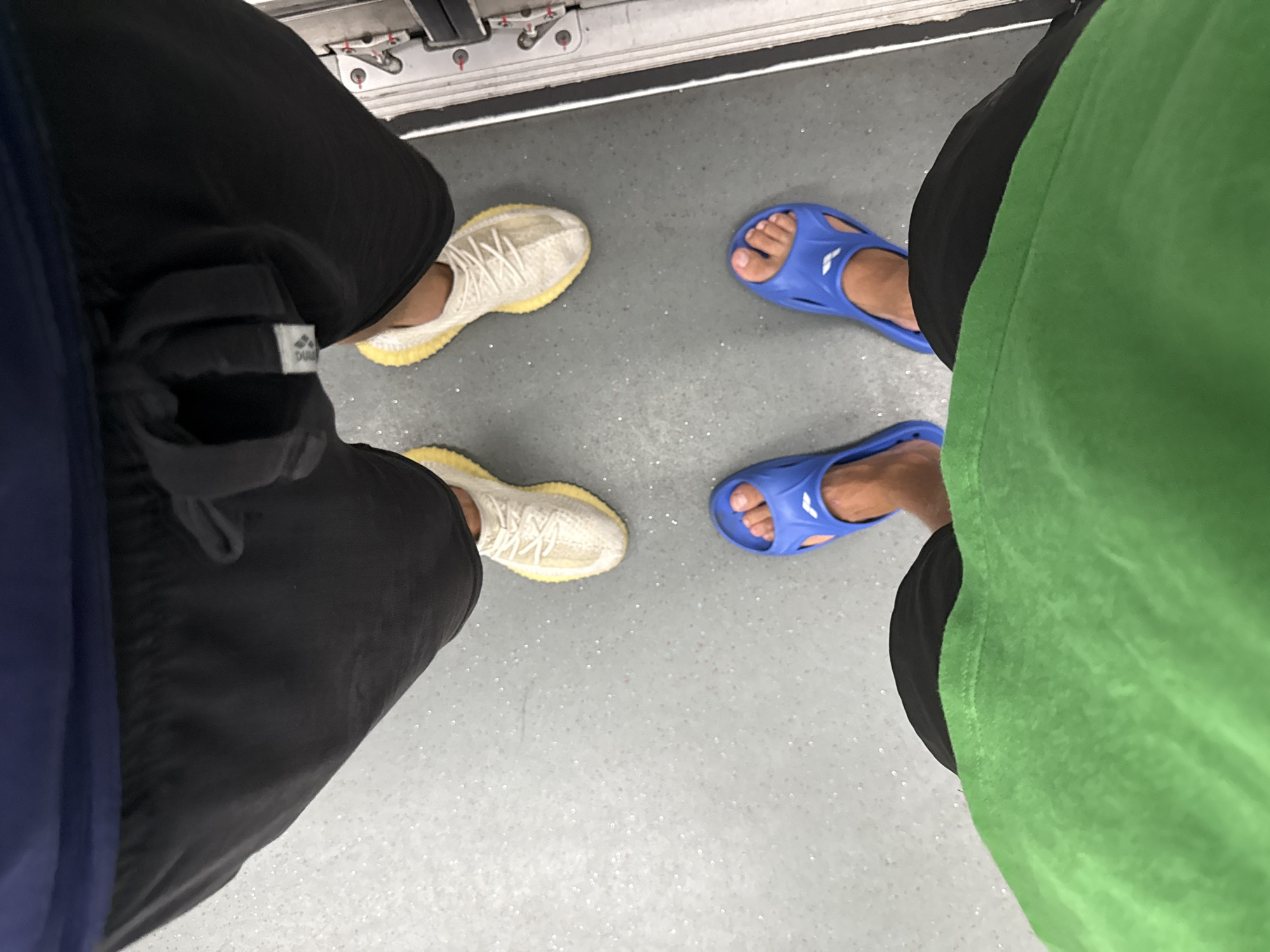 This high-angle shot depicts the lower bodies and feet of two individuals standing on a speckled gray floor, likely within a public transport vehicle in Tianhe District, China.
The person on the left is wearing black shorts and light-colored sneakers with a distinct ribbed, yellow sole, which appear to be Adidas Yeezy Boost 350 V2. A small, white fabric tag with partially legible text, possibly "DUA", is visible on an item attached to or part of their shorts. A dark blue garment is also visible on their left side.
The person on the right is wearing black shorts and a vibrant green shirt. They are wearing blue slip-on sandals, prominently featuring a white "ARENA" logo on the strap.
The floor surface is a light gray with numerous small, reflective speckles. At the top edge of the image, a metallic track or structural component, commonly found in subway or train cars, is visible. The image captures a static moment, focusing solely on the subjects' footwear and immediate surroundings. The indoor setting is evident, but no specific time of day, weather, or emotions are discernible.