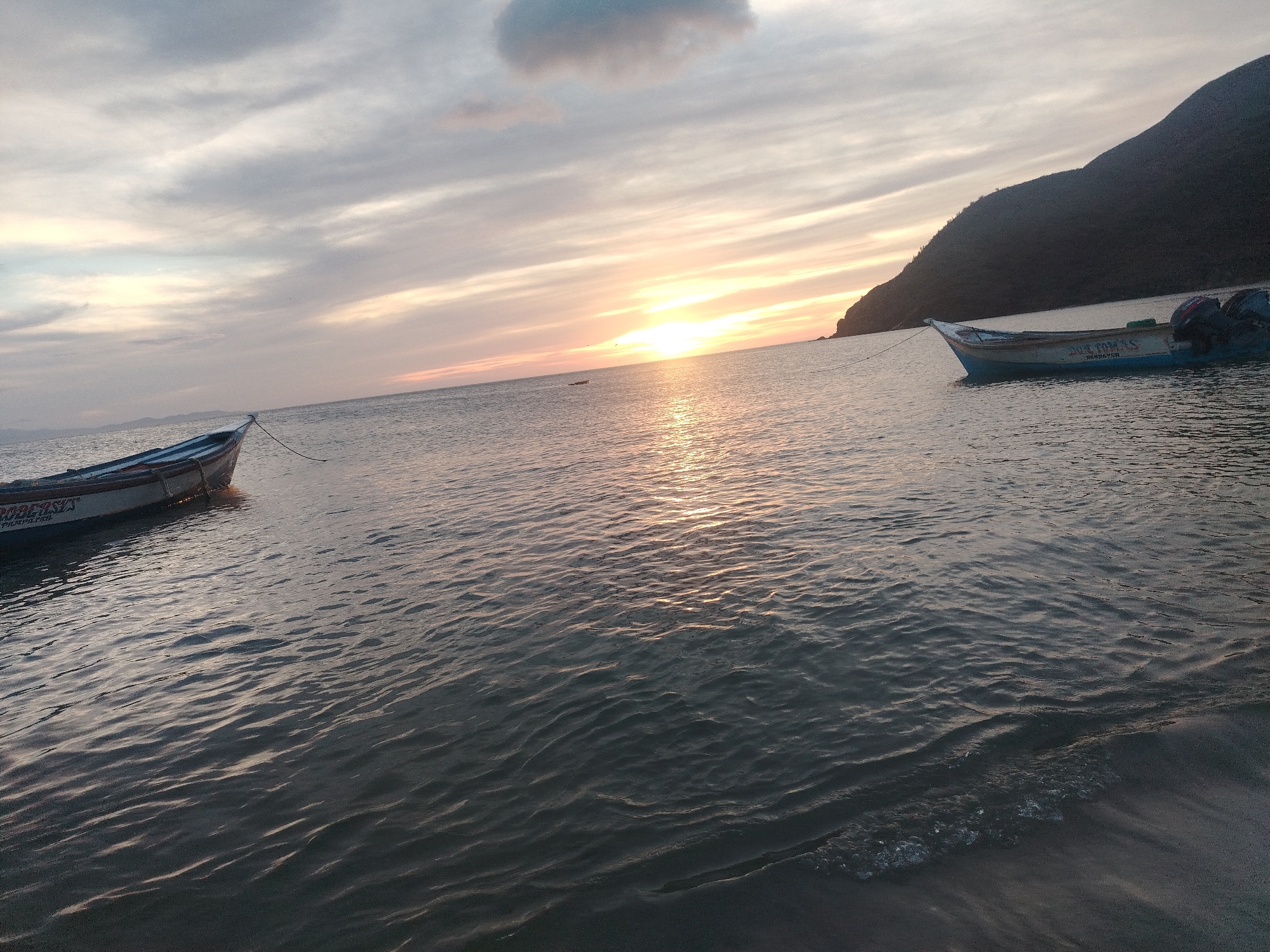 A serene seascape depicts a sunset over the ocean, with a gentle wave lapping at a sandy shore in the foreground. Two small fishing boats are anchored offshore, one on the left and another on the right, with a dark, mountainous silhouette rising in the background on the right side of the frame. The sky is a canvas of soft, wispy clouds in shades of gray, pink, and orange, with the sun casting a warm, golden glow on the water's surface. The overall ambiance is tranquil and picturesque.