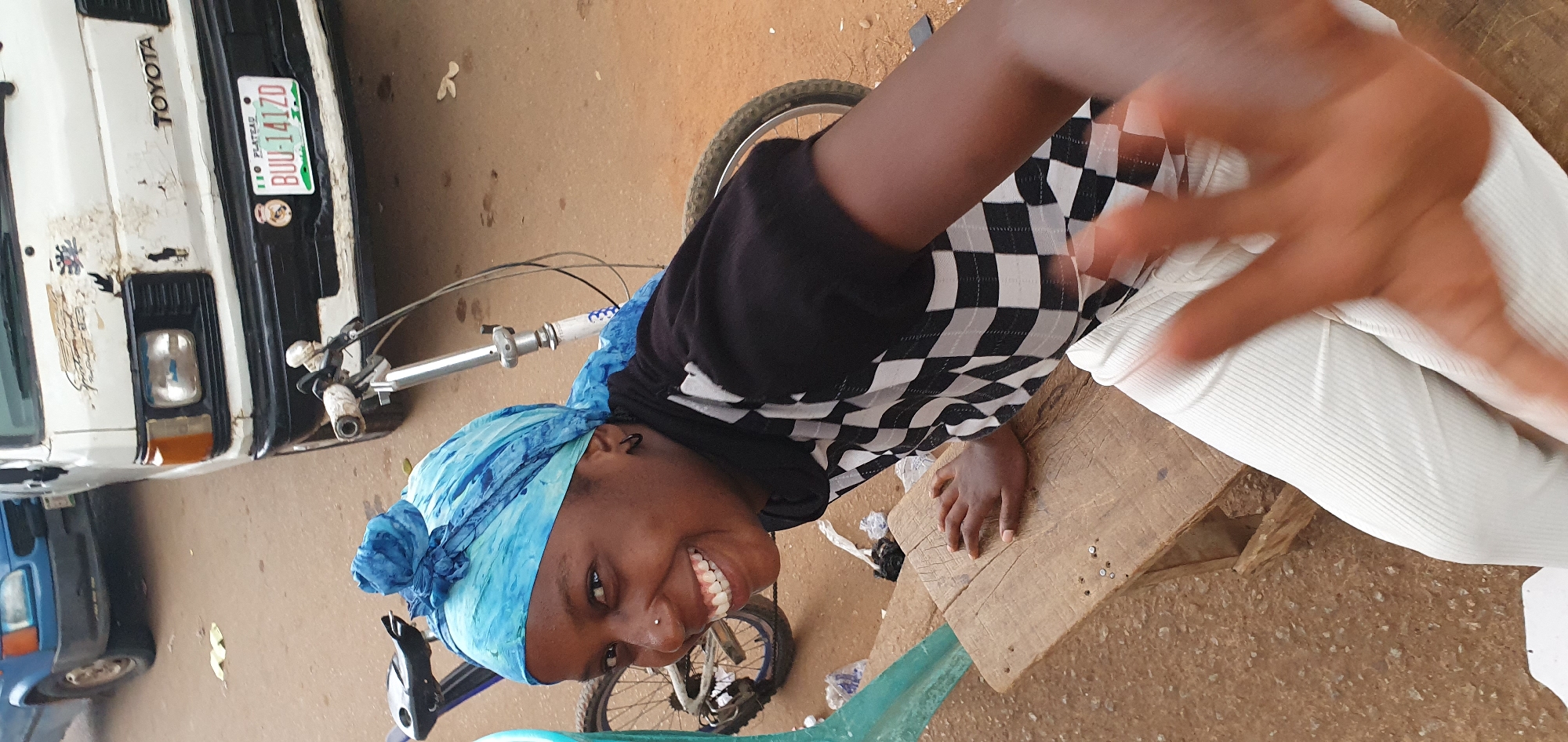 A young Black woman with a blue headscarf and a nose ring is smiling widely while sitting on a wooden bench. She is wearing a black and white checkered shirt and white pants. Her left hand is resting on the bench, and her right hand is blurred in the foreground, as if waving. A bicycle is partially visible behind her, and a white Toyota van with a Nigerian license plate is in the background. The scene appears to be outdoors on a dirt or paved surface, possibly in the city of Bukuru, Nigeria, based on the visible license plate which indicates "Plateau" state.