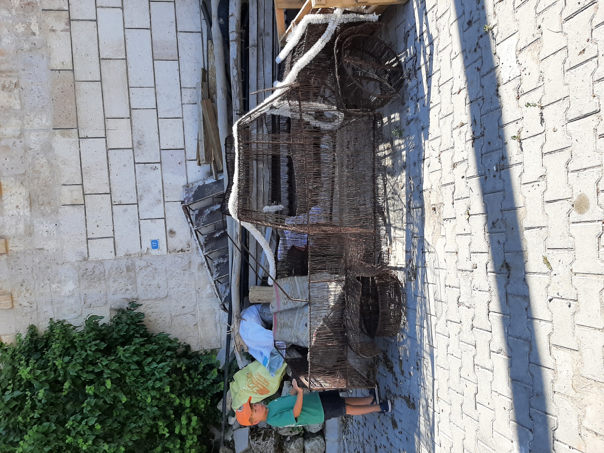 In Uçhisar Beldesi, Turkey, a young boy wearing an orange cap, green t-shirt, and dark shorts stands next to a large, rusty, wire-frame sculpture of a vehicle. The sculpture appears to be a repurposed item, possibly a large basket or cage, shaped like a car with its wheels visible. White fuzzy material is wrapped around parts of the sculpture, resembling decorative trim. Behind the sculpture, a weathered stone wall with a blue sign displaying the number "17" is visible. The ground is paved with interlocking concrete bricks. Various discarded items, including wooden planks, bags, and other debris, are piled up against the wall. Lush green foliage grows on the left side of the wall, partially obscuring it. The scene is bathed in sunlight, casting shadows on the ground.