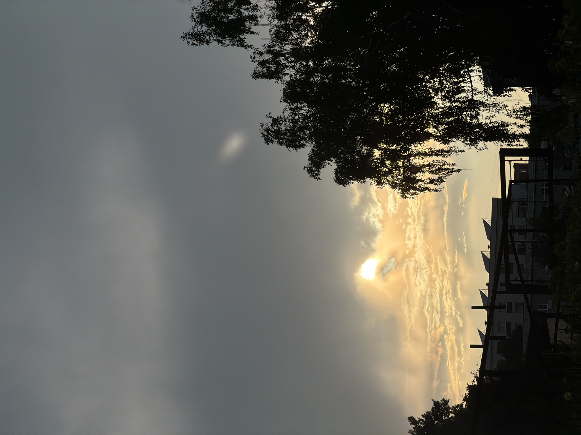The image depicts a cloudy sky at dusk, casting a subdued light over an urban scene. In the distance, a multi-story building with several solar panels on its roof is visible. The foreground is dominated by the dark silhouettes of trees. The sun is partially obscured by clouds, creating a bright, diffused glow in the sky. The scene appears to be an outdoor setting within the city of Sofia, Bulgaria.
