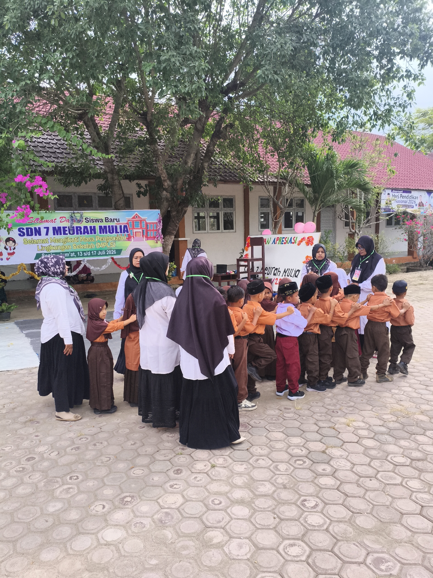 In front of a single-story building with red tile roofing, a group of children and adults are gathered on an outdoor paved area. The building appears to be a school, with windows visible along its facade. A large banner that reads "Selamat Datang Siswa Baru SDN 7 MEURAH MULIA" is displayed, indicating a welcome event for new students.
The group consists of approximately twenty children, mostly boys, dressed in uniform orange shirts and brown pants or skirts. They are arranged in a line, with their arms around each other's shoulders, suggesting they are participating in an activity or game. Several adult women, dressed in hijabs and modest clothing, are standing among the children, appearing to supervise or guide them. One woman in a white shirt and black hijab is facing the children, while another in a white shirt and black headscarf is standing to the side, holding a lanyard.
The environment is outdoors, with a large tree providing shade over part of the scene. Pink flowers are visible on a vine to the left of the frame, and some greenery and palm fronds are present. The paving stones are hexagonal and have a textured surface. The overall atmosphere suggests a school event, possibly the beginning of a new academic year. The presence of balloons and streamers adds a festive touch. The scene is well-lit, indicating daylight. No specific details about the city of Baratpayaitek, Indonesia are present in the image itself, beyond what is written on the banner.