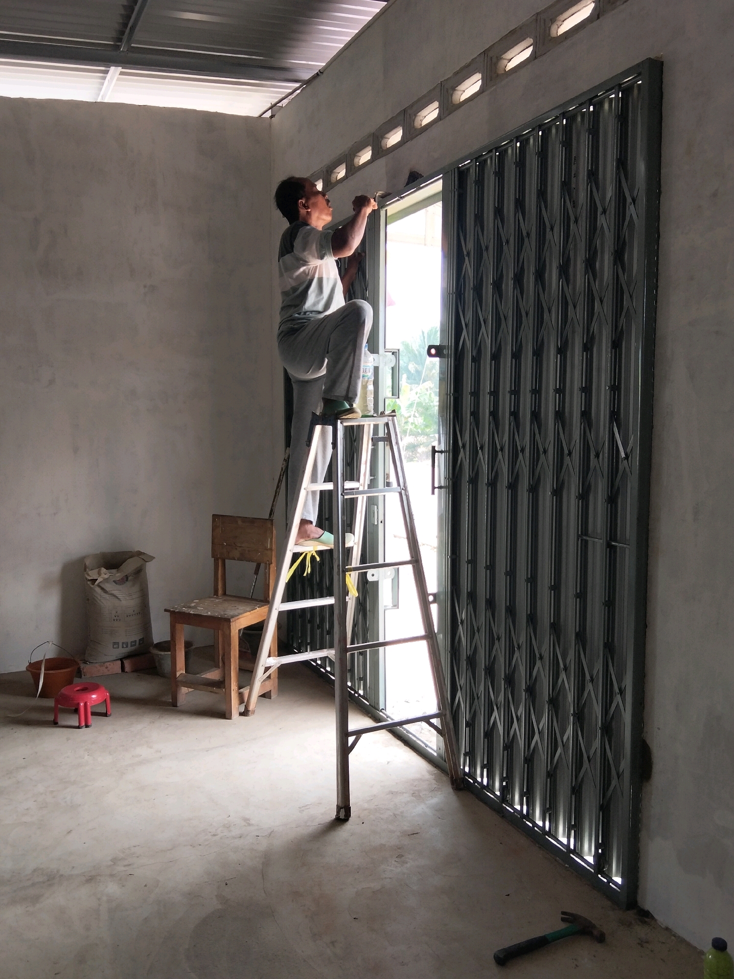 The image depicts a scene inside a building under construction, likely in Perambahan, Indonesia. A person is standing on a silver ladder, positioned in front of a metal accordion gate that covers an opening. The person appears to be working on the top part of the gate, possibly installing or adjusting it. They are wearing a striped t-shirt and light-colored pants.
The environment is a concrete-floored room with unfinished walls. To the left of the ladder, there is a small wooden chair, a bag of cement, a red plastic stool, and some bricks. A hammer lies on the floor near the bottom right of the image. Natural light is entering from the open doorway behind the gate, and the ceiling appears to be made of corrugated metal. The overall impression is of an active construction or renovation site.