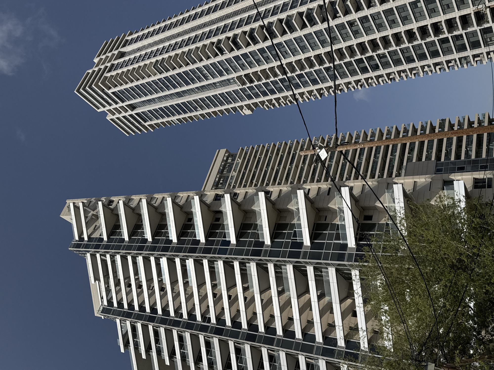 The image, taken from a low angle, depicts two buildings in Tel-Aviv, Israel, against a clear blue sky. The architectural style is modern, with sharp lines and repetitive geometric patterns. The building in the upper portion of the frame features a series of cantilevered balconies, creating a stepped effect. The building below has vertical white lines that run down the surface of the building. Power lines run diagonally across the frame, and the top branches of a tree are visible on the right side. The overall impression is one of urban density and contemporary design.