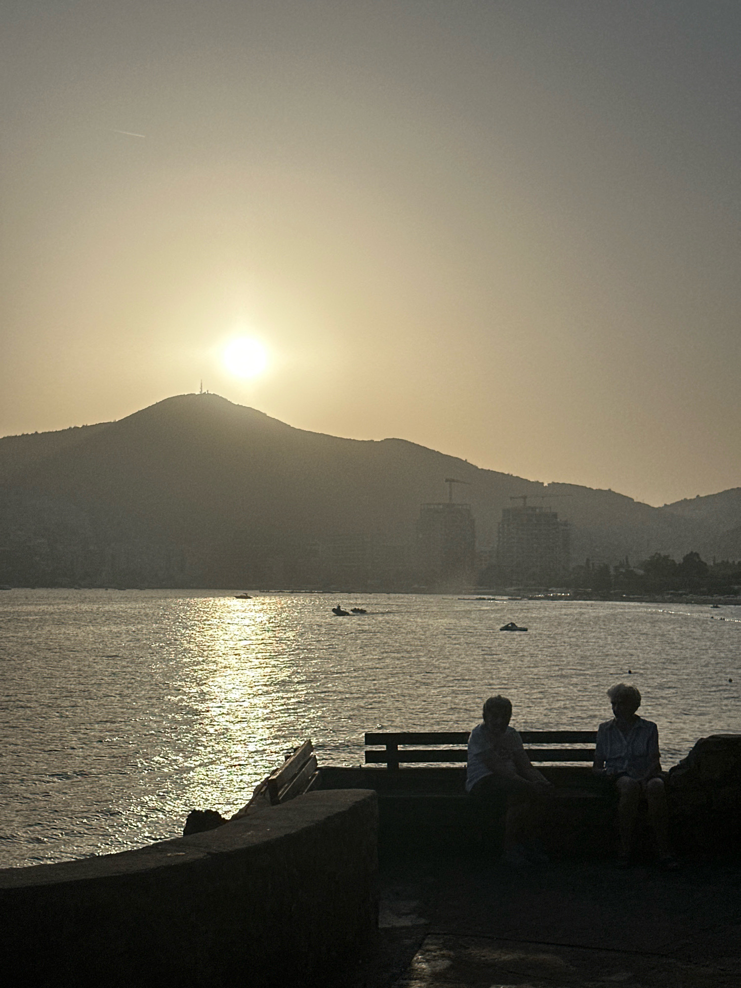 The image depicts a serene sunset over the Adriatic Sea in Budva, Montenegro. The sun, a bright orb partially obscured by atmospheric haze, casts a golden glow across the water, creating a shimmering path of light. In the foreground, two elderly women are seated on a dark bench, silhouetted against the bright sea and sky. They appear to be conversing or enjoying the view. Behind them, the water is dotted with several small boats and a jet ski. Further in the background, a large, mountainous island dominates the horizon, topped with a communications tower. The coastline is dotted with buildings, some of which appear to be under construction, indicated by visible cranes. The overall atmosphere is peaceful and tranquil, with the golden light of the setting sun bathing the scene in a warm, diffused light. The vantage point appears to be from a promenade or a pier overlooking the bay.