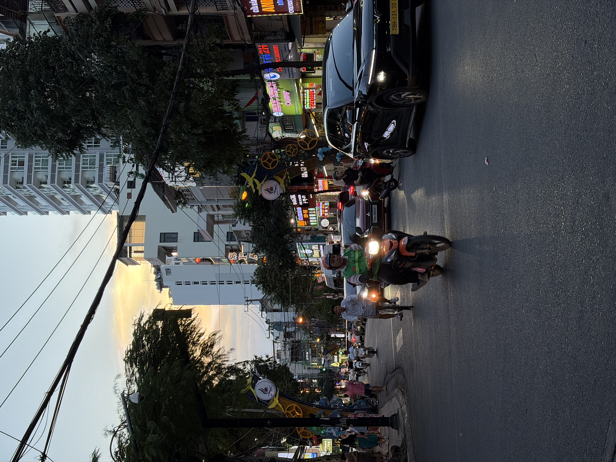 This image captures a bustling street scene in Nha Trang Ward, Vietnam, likely during the early evening as indicated by the warm glow of the setting sun and the illuminated neon signs. The street is filled with activity, with numerous motorbikes and a few cars navigating the asphalt road. People are seen riding motorbikes, walking on the sidewalks, and engaged in various activities.
The setting is urban, characterized by multi-story buildings lining the street, many of which are adorned with colorful neon signs advertising businesses. Some of these signs display text in Vietnamese, such as "CỬA HÀNG" (store) and "IS LAUNDRY." The presence of trees adds a touch of greenery to the urban landscape, contrasting with the man-made structures. Electrical wires are visibly strung across the scene, typical of many streets in Southeast Asia.
Notable details include the bright headlights of vehicles, suggesting it's either dusk or a slightly overcast day. The overall atmosphere appears dynamic and lively, a common characteristic of busy city streets in Vietnam, especially as evening approaches and businesses begin to glow under artificial light. The image is oriented in a way that suggests it might have been captured by someone holding their phone vertically.