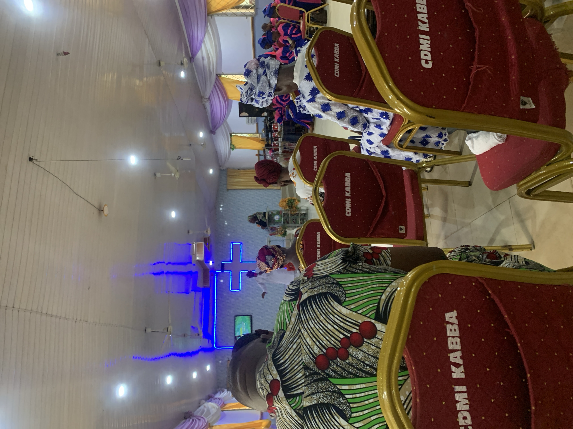The image captures an indoor scene in what appears to be a religious gathering, possibly a church service, within the city of Kabba, Nigeria. Occupying the foreground are several red and gold chairs, prominently displaying the inscription "CDMI KABBA" on their backrests. These chairs are arranged in rows, suggesting a formal seating arrangement for attendees. Several people are seated, their attire reflecting traditional Nigerian clothing styles, including vibrant colors and patterned fabrics. Notably, some women are wearing headwraps (gele). In the background, a blue neon cross is illuminated on the wall, a symbol often associated with Christianity. The environment is well-lit, with a high ceiling. The interaction is of people at a religious service.