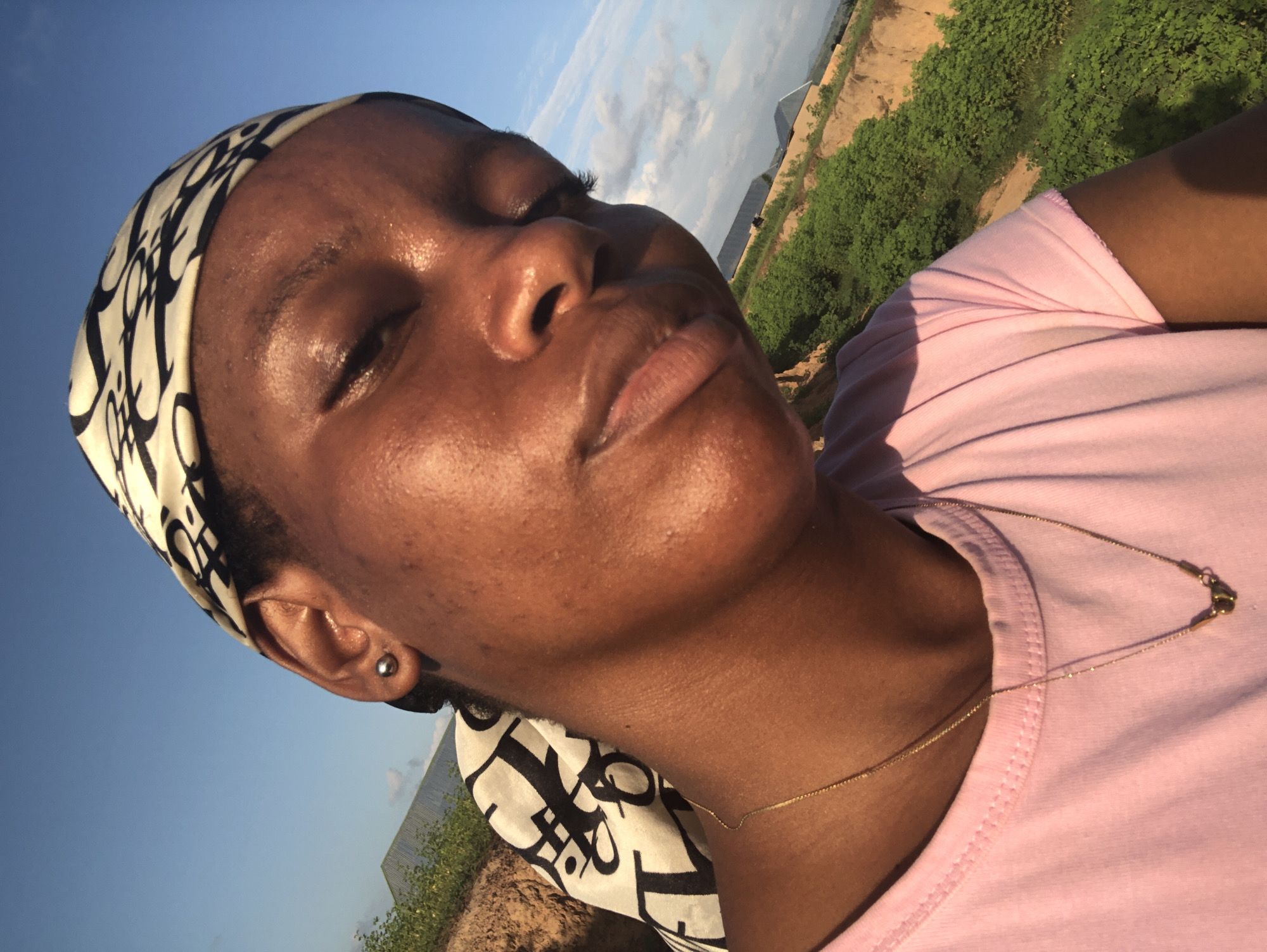 A close-up selfie of a young Black woman wearing a pink t-shirt and a black and white patterned headscarf. She has a silver stud earring in her visible left ear and a thin gold necklace with a small pendant. The woman is outdoors, with a clear blue sky with some clouds in the background. Lush green vegetation and some buildings are visible on a hill in the distance, suggesting a rural or semi-rural setting. The lighting indicates it is daytime, likely late afternoon due to the warm glow on her skin. The image appears to have been taken in Kashere, Nigeria, given the provided context.