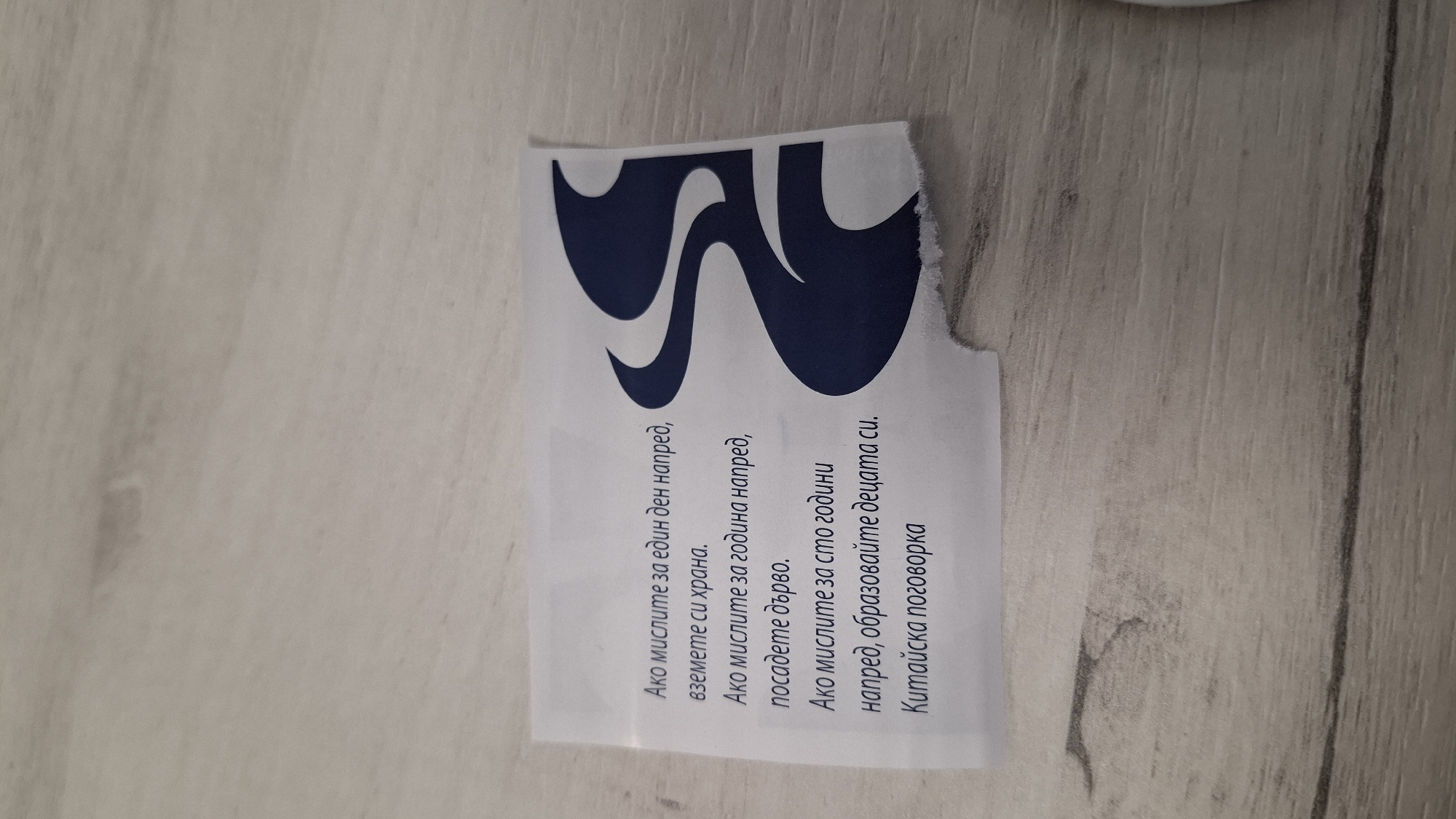 A torn piece of white paper with black text and a blue stylized graphic is lying on a light-colored wooden surface. The text, which appears to be a proverb, is in Bulgarian. The graphic is on the right side of the paper and features a swirling design. The wooden surface has a visible grain pattern and some slight discoloration. The lighting is even, casting no harsh shadows. There are no people or identifiable locations visible in the image.