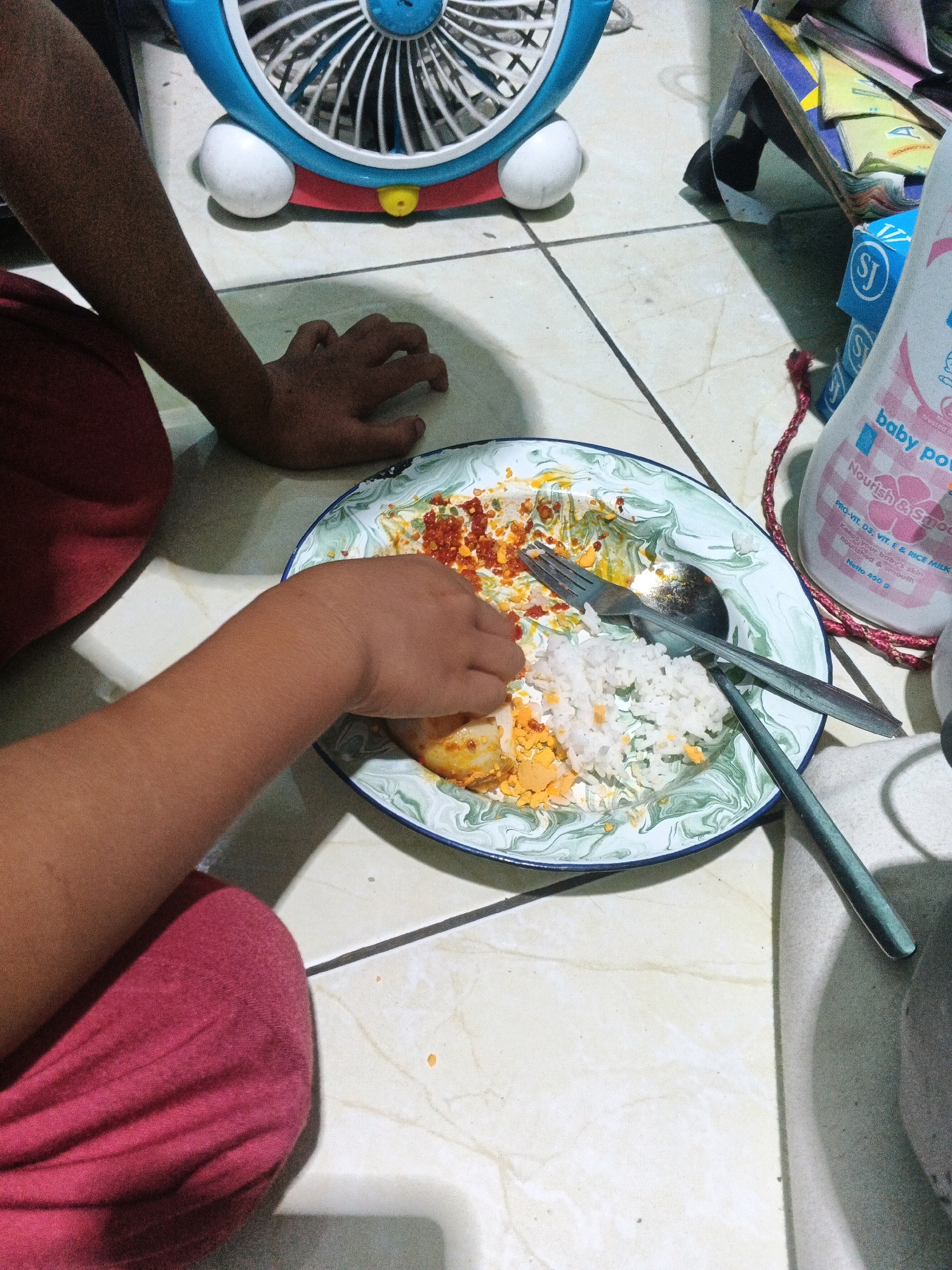A high-angle, medium shot captures a child in a red garment sitting on the floor in what appears to be a residential setting in Kadu, Indonesia, engaged in eating from a plate of food. The child's left arm and hand are actively reaching into the plate, which contains rice, what looks like chopped hard-boiled egg, and chili flakes, all spread across a decorative ceramic plate with a green marble pattern. A fork and spoon are also present on the plate, partially submerged in the food. To the left of the plate, the child's right arm and hand are visible, resting on the floor. In the background, a blue and white fan with a red base is partially visible, along with stacks of colorful papers or books, and a white bottle of baby powder with pink lettering. The floor is tiled with a light-colored marble pattern. The lighting suggests a well-lit indoor environment, casting shadows on the floor.