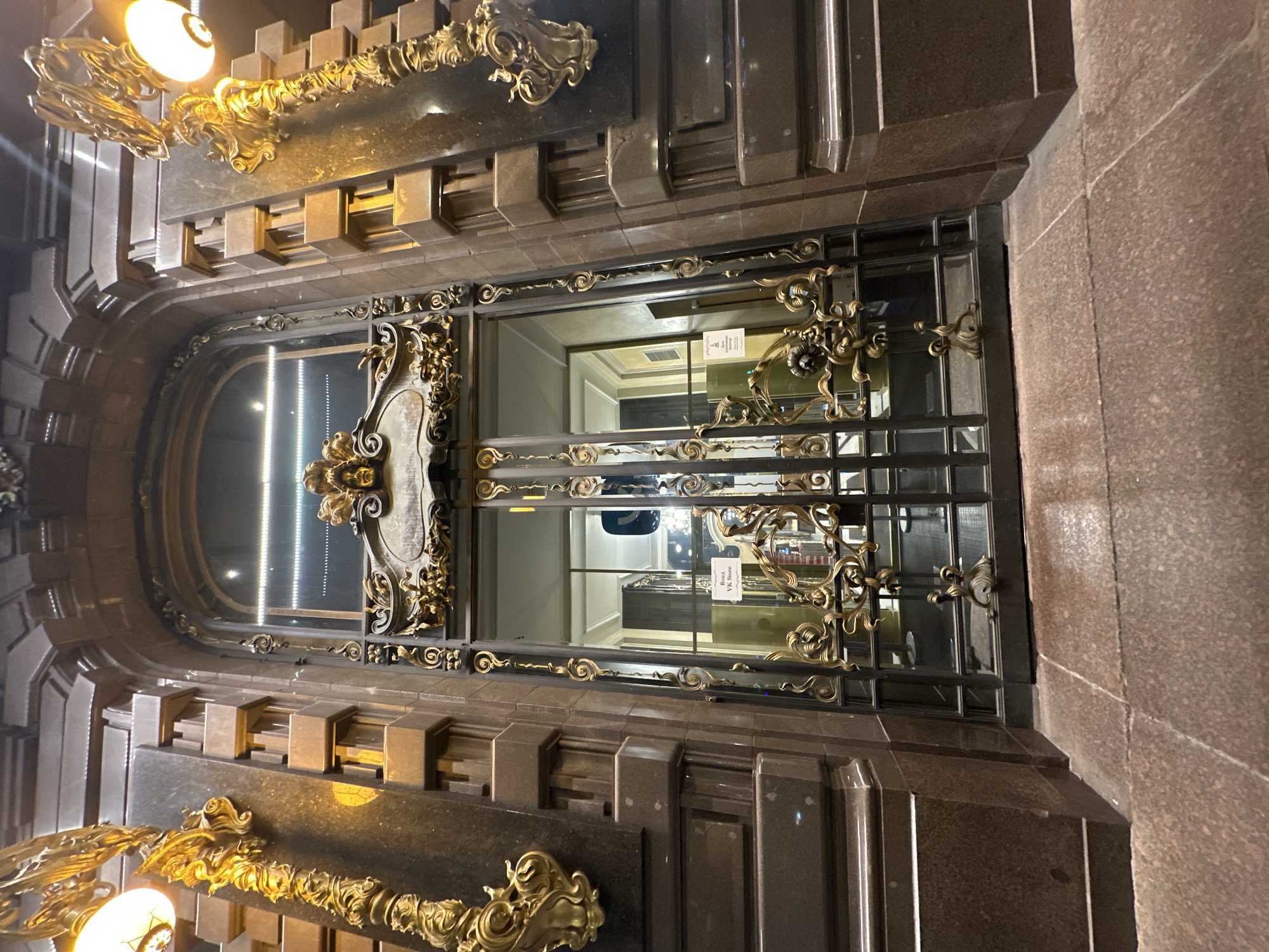 The image shows the entrance to the BURO V6 Store in Saint Petersburg, Russia. The entrance is adorned with elaborate, ornate gold detailing around the doorway and on the security gate. The door itself is constructed of a dark metal frame. The building is made of dark stone. A light fixture is mounted on the upper left. The interior of the store is visible through the gate.