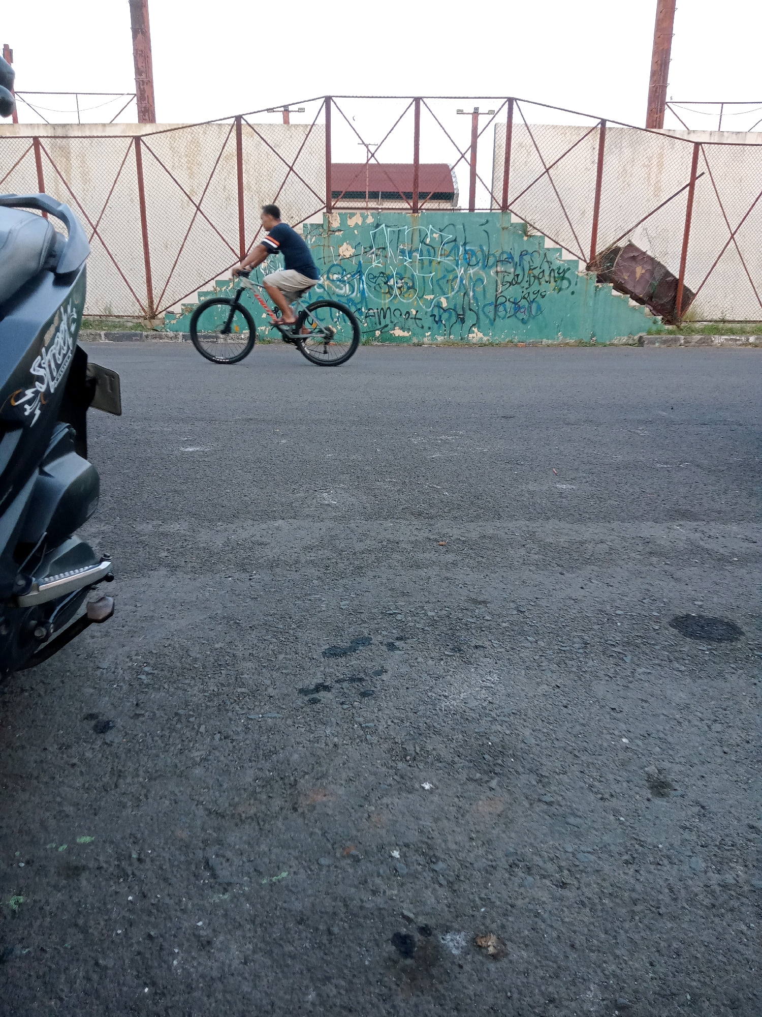 A person is riding a bicycle on a paved road in front of a graffiti-covered wall. The cyclist is wearing a dark blue t-shirt and light-colored shorts. A motorcycle is parked in the foreground on the left side of the image, partially obscuring the view of the road. In the background, there is a chain-link fence with a rusted metal structure. The sky is overcast. The scene appears to be in an urban or semi-urban setting in Karawang, Indonesia.