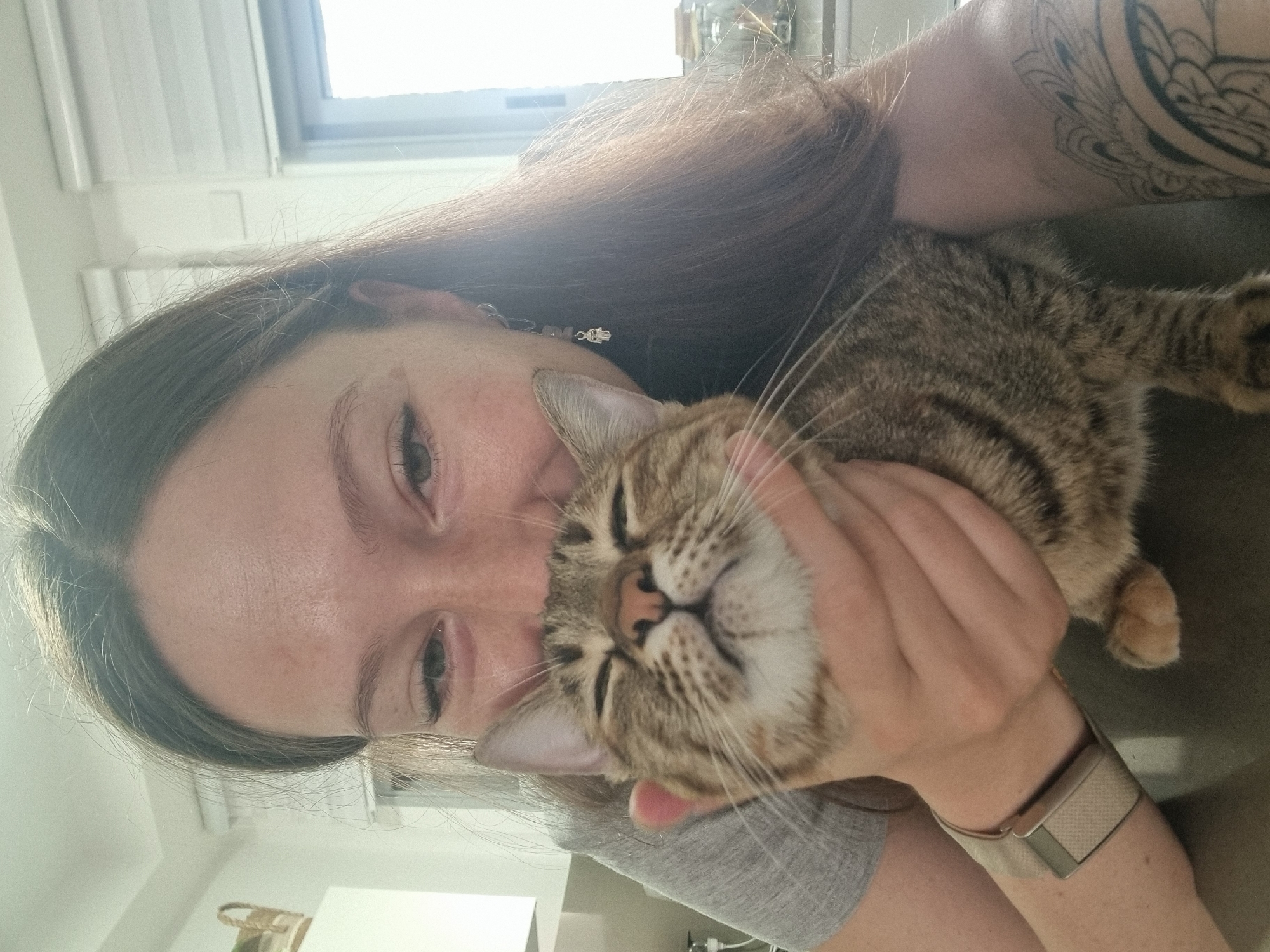 A woman is holding a tabby cat, pressing its face against hers. The woman has long brown hair, fair skin, and is wearing a gray shirt and a watch with a light-colored band. Part of a tattoo is visible on her upper arm. The cat appears relaxed, with its eyes closed. The background includes a window with blinds and a glimpse of the interior of a room. The location is not discernible as being in Ramat Gan, Israel, from the image.