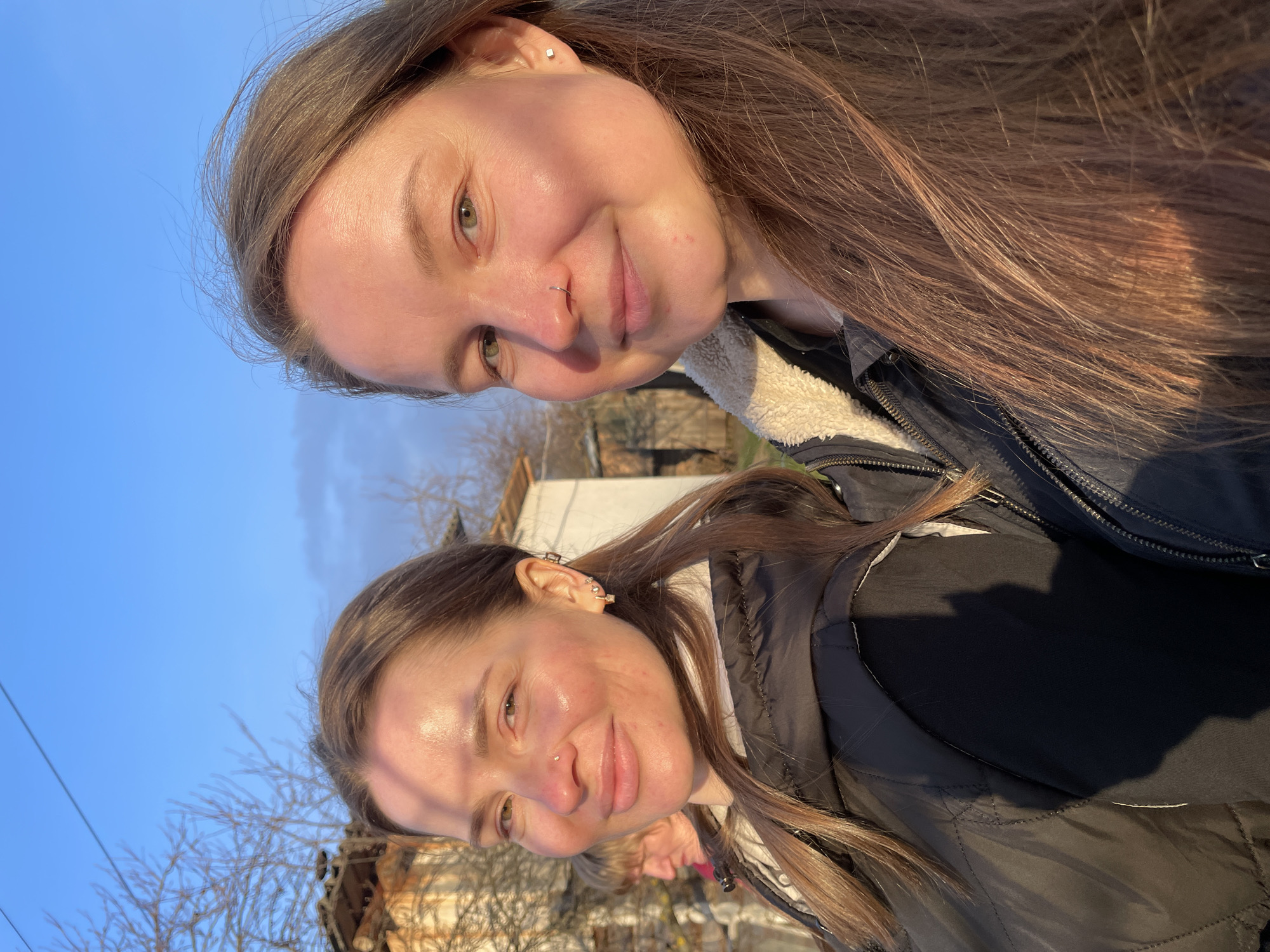 This outdoor portrait features two young women captured in a close-up, likely a selfie, under the warm glow of late afternoon or early evening sunlight. A third person's head is partially visible in the distant background. The scene is set against a clear blue sky with bare trees, suggesting a colder season, in what appears to be a rural or suburban area of Балдейка, Russia.
The woman positioned closer to the viewer (top in the original image orientation) has long, straight brown hair. She has light-colored eyes, a gentle smile, and wears a small silver hoop nose ring on her right nostril. A subtle square stud piercing is visible on her left earlobe. She is dressed in a dark jacket, possibly a puffer style, with a cream or white shearling-like lining visible at the collar.
The second woman, slightly behind and below the first, also has long brown hair and a soft smile. She has multiple piercings on her left ear, including a stud on the lobe and several decorative helix or cartilage piercings. A tiny, almost unnoticeable stud is present on her right nostril. She is wearing a black puffer jacket over a lighter-colored garment.
The sunlight bathes their faces and hair, creating highlights and a warm, pleasant atmosphere. In the background, out-of-focus residential buildings and the stark outlines of bare tree branches are visible against the vibrant blue sky. No visible text is present in the image.