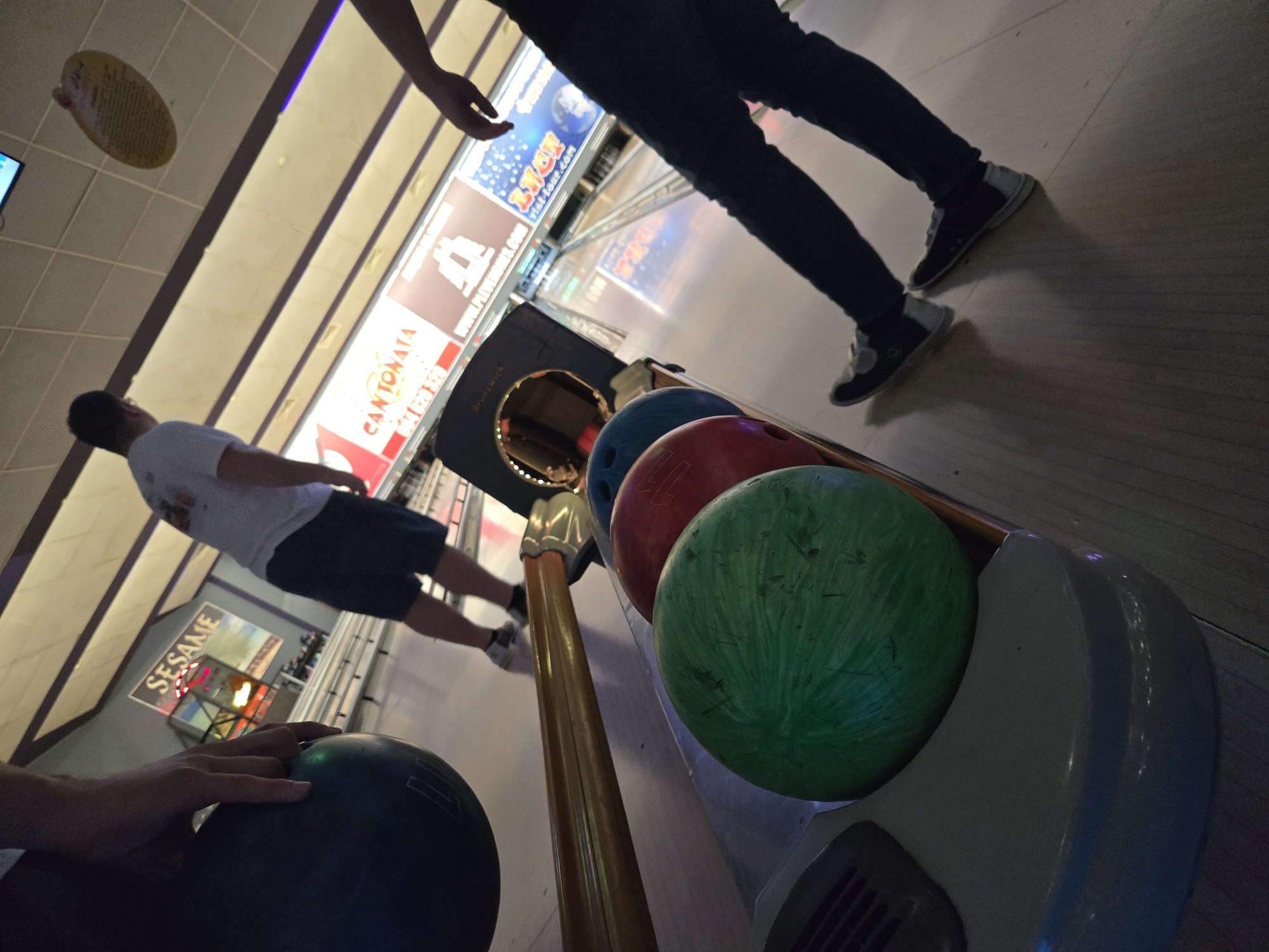The image shows an indoor bowling alley. There are multiple bowling balls of different colors (green, red, blue, and black) sitting in a ball return. Two people are visible, one in a white t-shirt and dark shorts standing on the lane, preparing to bowl, and another standing further down the lane, wearing jeans and black/grey shoes. A third person's hand is on a bowling ball. In the background are illuminated signs and screens above the bowling lanes. One sign reads "SESAME." Another sign reads "CANTONATA."
I cannot confirm the location in the city of Pleven, Bulgaria, from the image content alone.
