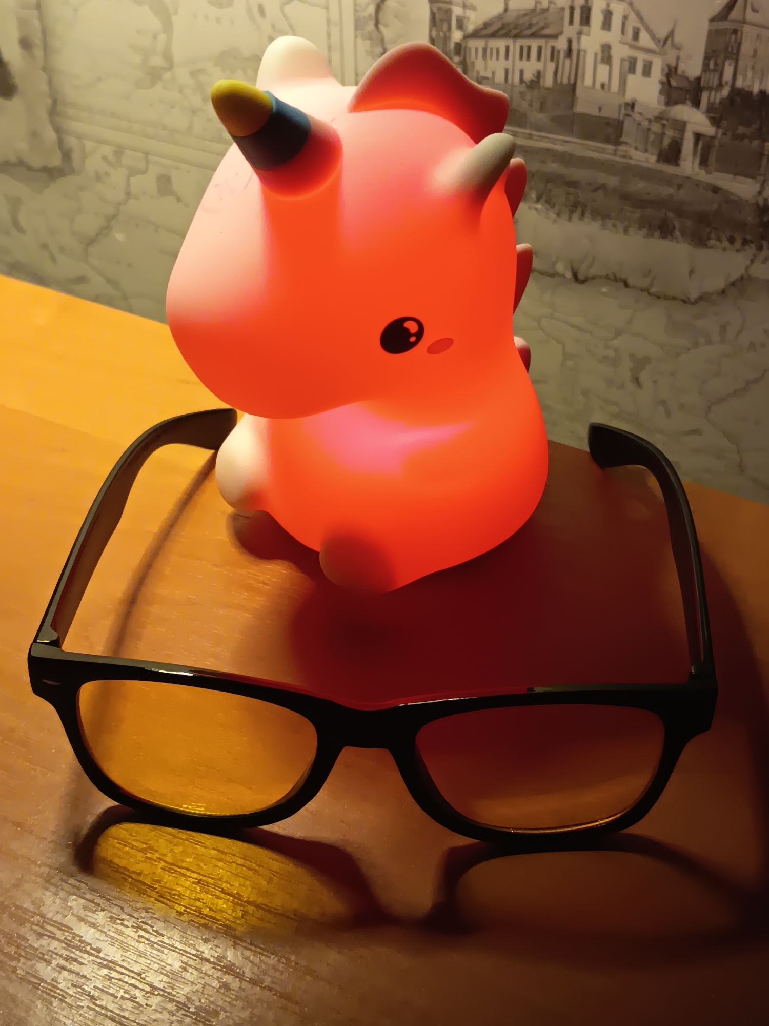 The image shows a close-up of a pink, illuminated unicorn-like toy sitting on a wooden table, with a pair of black-framed glasses in front of it. The toy is glowing with an orange light and has a single horn with blue and yellow accents. The background features a black and white mural or wallpaper depicting buildings and a body of water. There are no people visible in the image. The scene is lit with artificial light, casting shadows on the table. The location is Izmail, Ukraine.