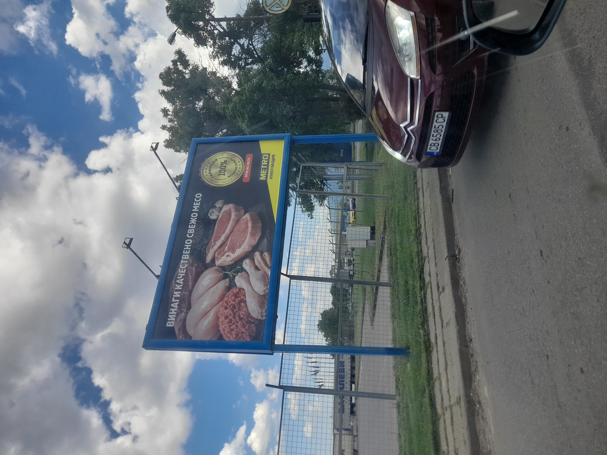 The photograph captures a roadside scene in Voluyak, Bulgaria. A billboard promoting "METRO Живей На Едро" (METRO Live Big) and advertising fresh meat is prominently displayed, featuring images of various cuts of meat. The sign also boasts a "100%" guarantee with a money-back pledge. A maroon car, partially visible, is parked adjacent to the billboard, with its license plate reading "CB 6585 CP." The environment includes a grassy area, a chain-link fence, and trees in the background, partially obscured by the billboard. The sky is visible above the billboard, exhibiting a mix of blue and white clouds.
