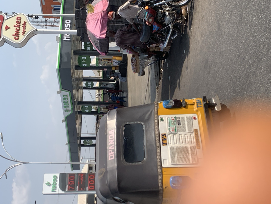 The image captures a daytime street scene in Lokoja, Nigeria, centered around a Habso Filling Station and an adjacent Chicken Republic.
In the foreground, a yellow Keke Napep (tricycle) is positioned with its rear facing the viewer. The tricycle features a dark canopy with "OPANDA" printed in pink text. Its rear panel displays "TVS" branding, a Nigerian license plate "KUE-866VZ," and a contact number "07019100360." The lower-left portion of the image is partially obscured by an orange blur. The tricycle rests on a paved asphalt road.
To the right of the tricycle, near the roadside, a street vendor operates under a pink and white striped umbrella. A cart filled with produce, possibly onions or garlic, is visible under the umbrella. Two adult males are present near this stall: one in a striped shirt faces another man in a light-colored top and a cap, who stands next to a red and black motorcycle parked on its kickstand. A second, darker motorcycle is partially visible behind the vendor's cart.
The midground is dominated by the Habso Filling Station, identifiable by its prominent green and white signage and a large canopy covering multiple fuel pumps. Price displays on a tall Habso sign to the left show "PETROL 790," "DIESEL --WE," and "KERO 000" (partially visible). Pump numbers such as "3/5," "4/3," "12/11," "2/1," and "10/9" are visible under the canopy. Several individuals are present within the filling station area.
Above the filling station canopy, to the right, a "Chicken Republic" sign on a tall pole is clearly visible. The sky is bright and clear, indicative of fair weather. A multi-lamp street light pole extends into the upper left quadrant of the frame.