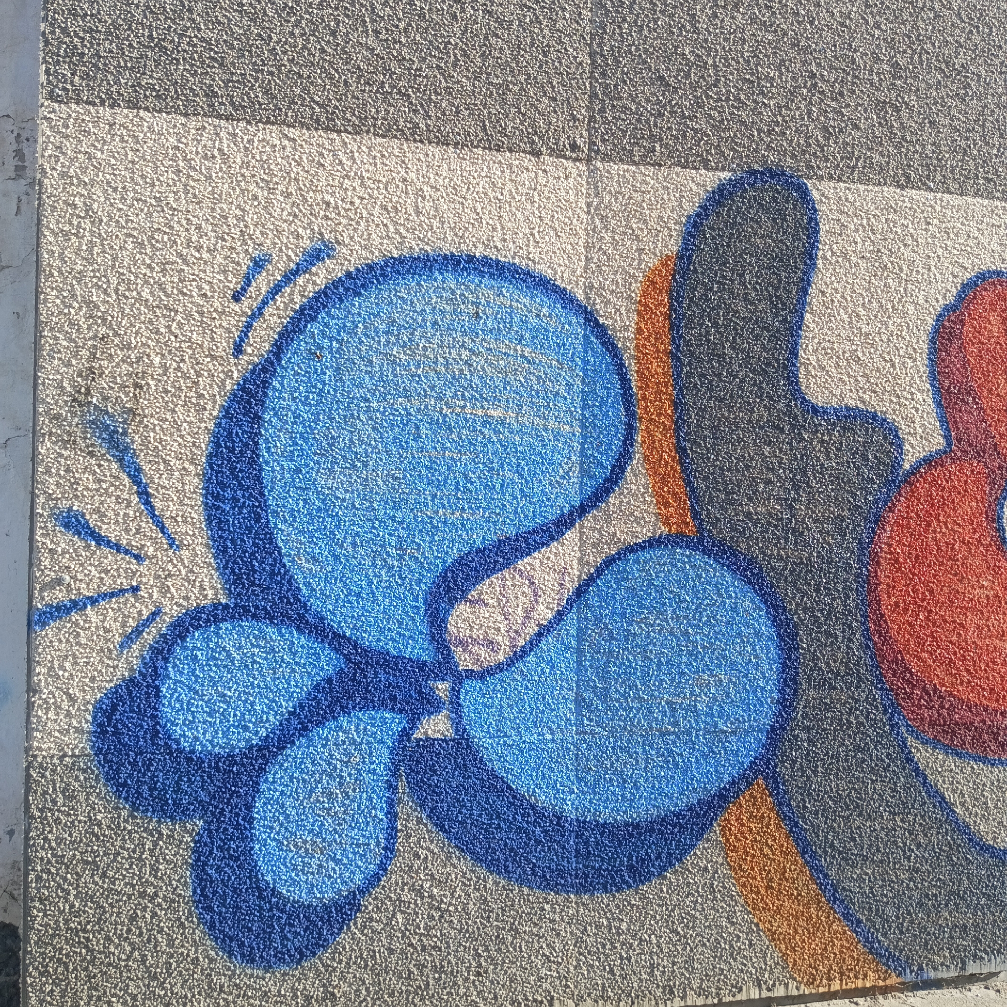 The image shows a section of a wall with graffiti art. The background is a light-colored, textured surface. The graffiti consists of stylized shapes, predominantly in blue, black, orange, and red. The blue section features a larger circular form connected to smaller shapes resembling droplets. There are radiating lines emanating from the left side of the blue shape, suggesting motion or energy. The other shapes are abstract, with outlines in a darker shade of blue. The wall appears to be composed of panels, indicated by the change in texture at the top of the frame.
This image was captured in the city of Chaikovsky, Russia.